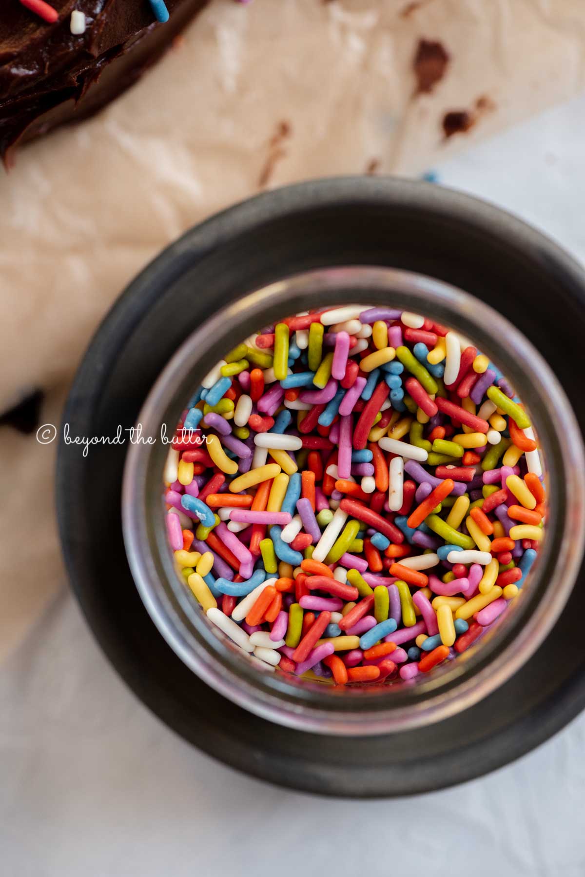 Small bowl of rainbow sprinkles on a dark plate with chocolate cake next to it | All Images © Luyiz®