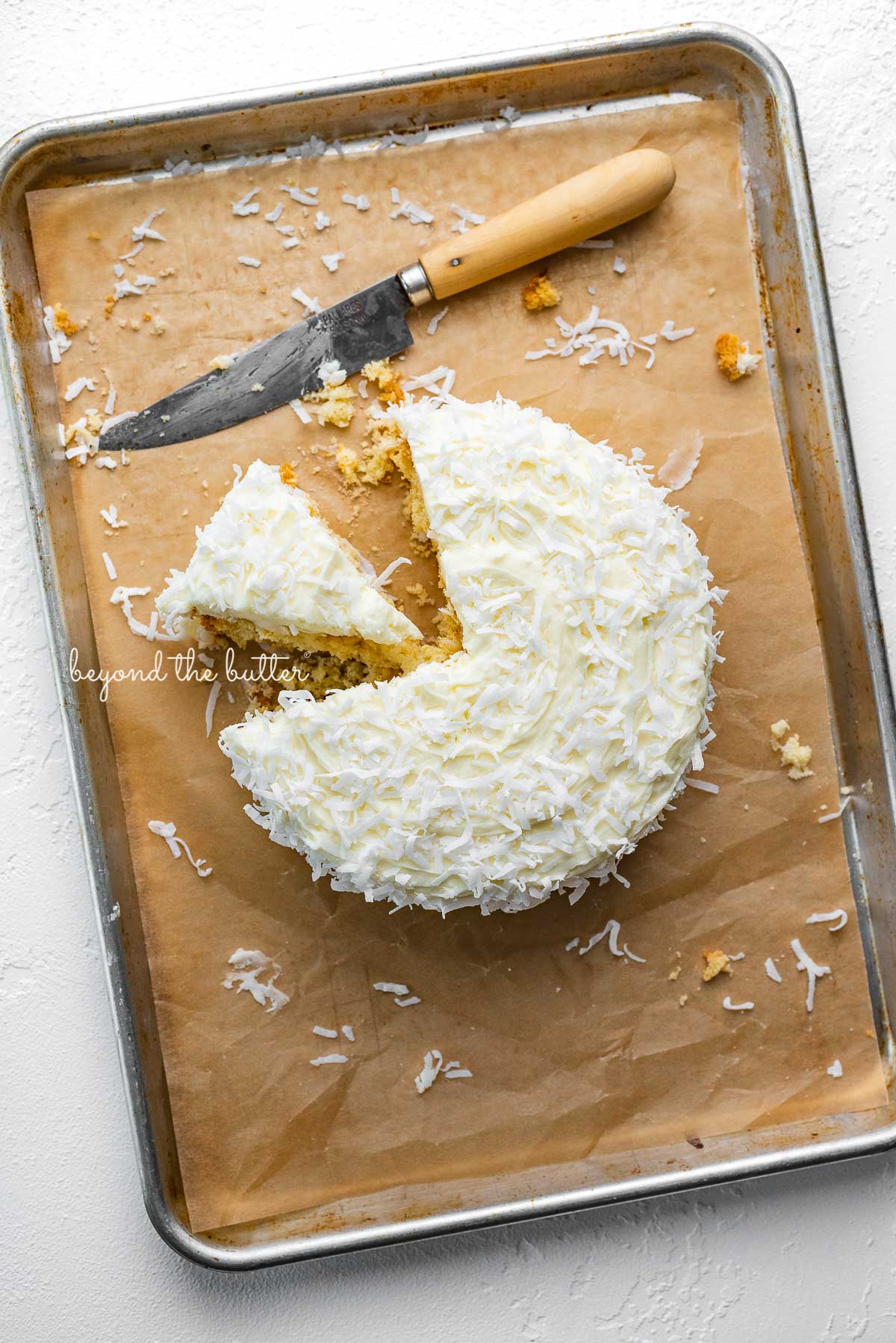 Single layer coconut cake frosted with coconut buttercream with slice cut out on natural parchment paper-lined baking sheet | © Luyiz®