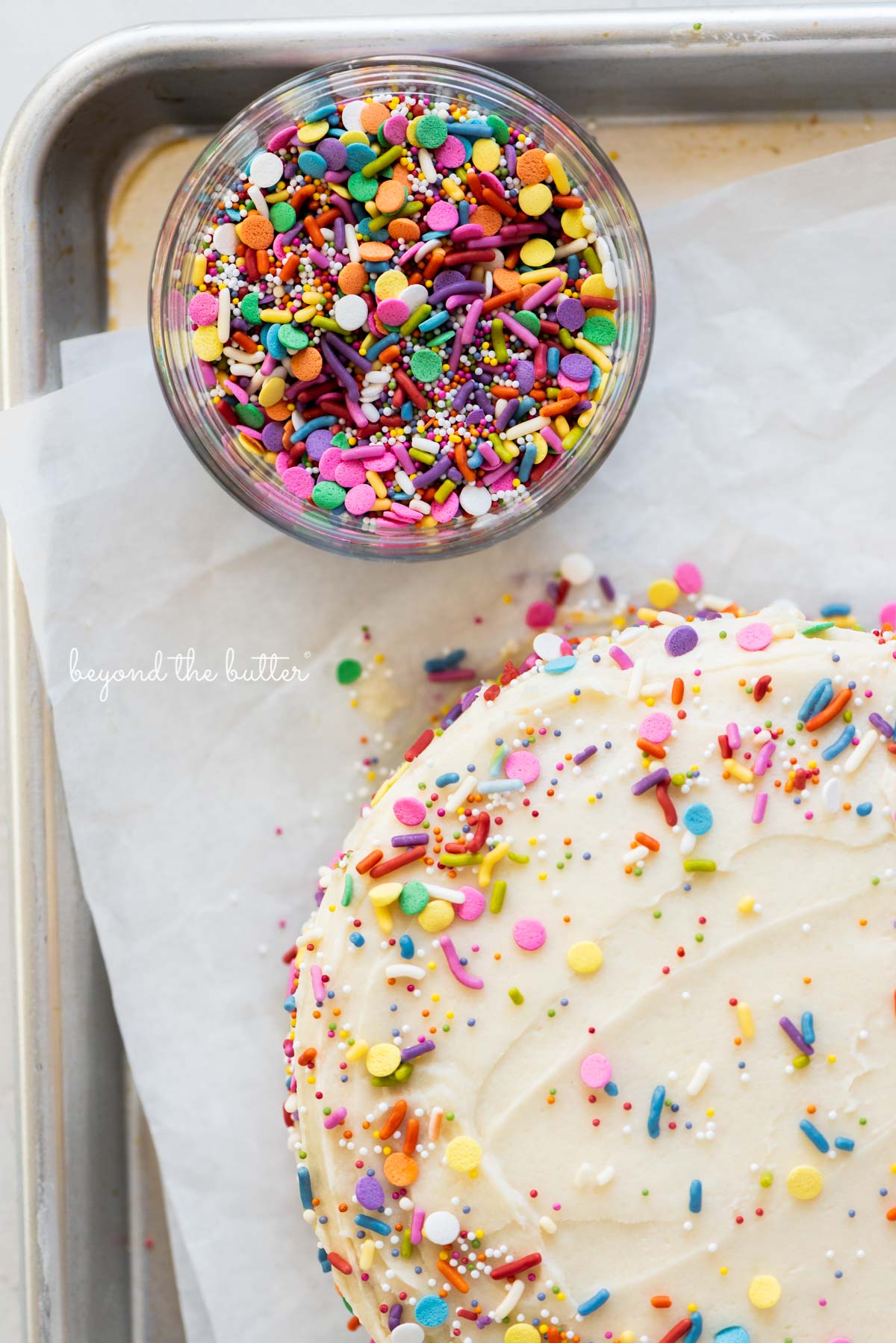 Close up image of rainbow jimmies and single layer funfetti cake on a parchment paper lined baking sheet from Luyiz | All images © Luyiz®