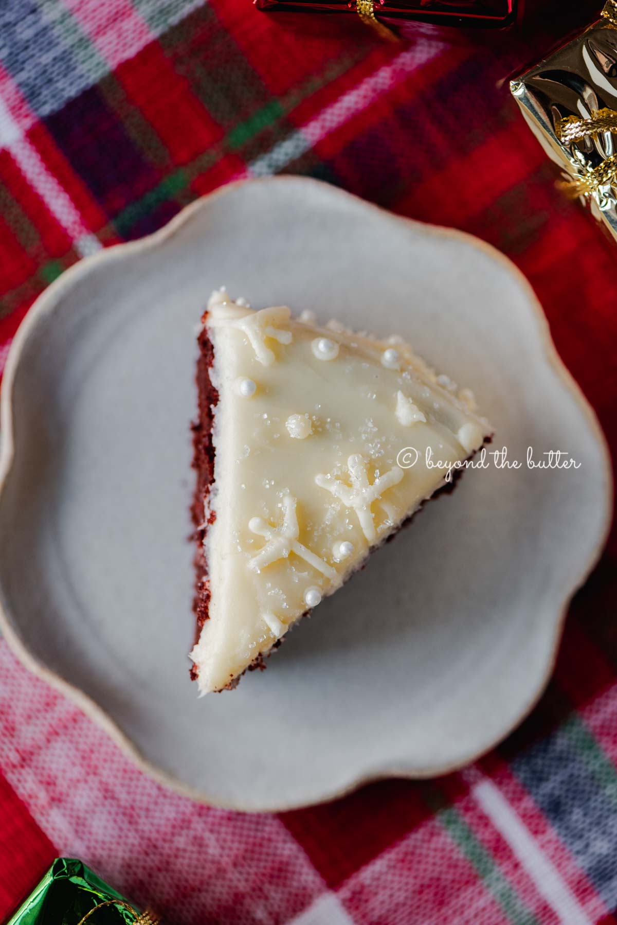 Slice of red velvet cake with cream cheese frosting on a scalloped edge dessert plate with red plaid background | All Images © Luyiz®