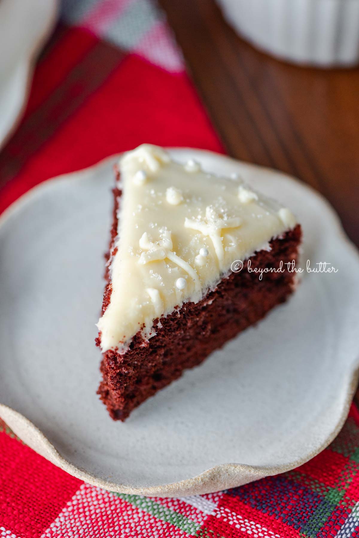 Slice of red velvet cake with cream cheese frosting on a scalloped edge dessert plate with red plaid background | All Images © Luyiz®