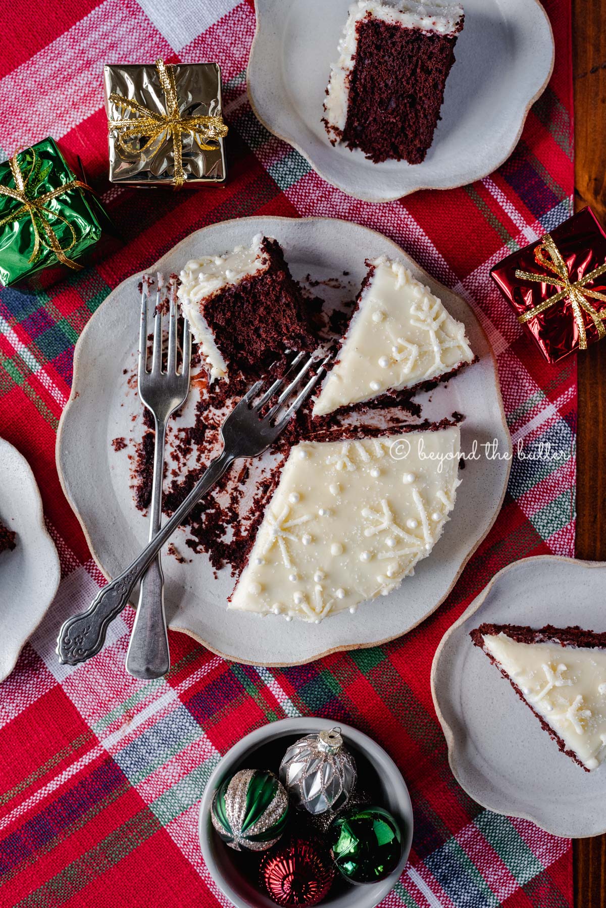 Half eaten and sliced red velvet cake with cream cheese frosting on a scalloped edge dessert plate with red plaid background | All Images © Luyiz®