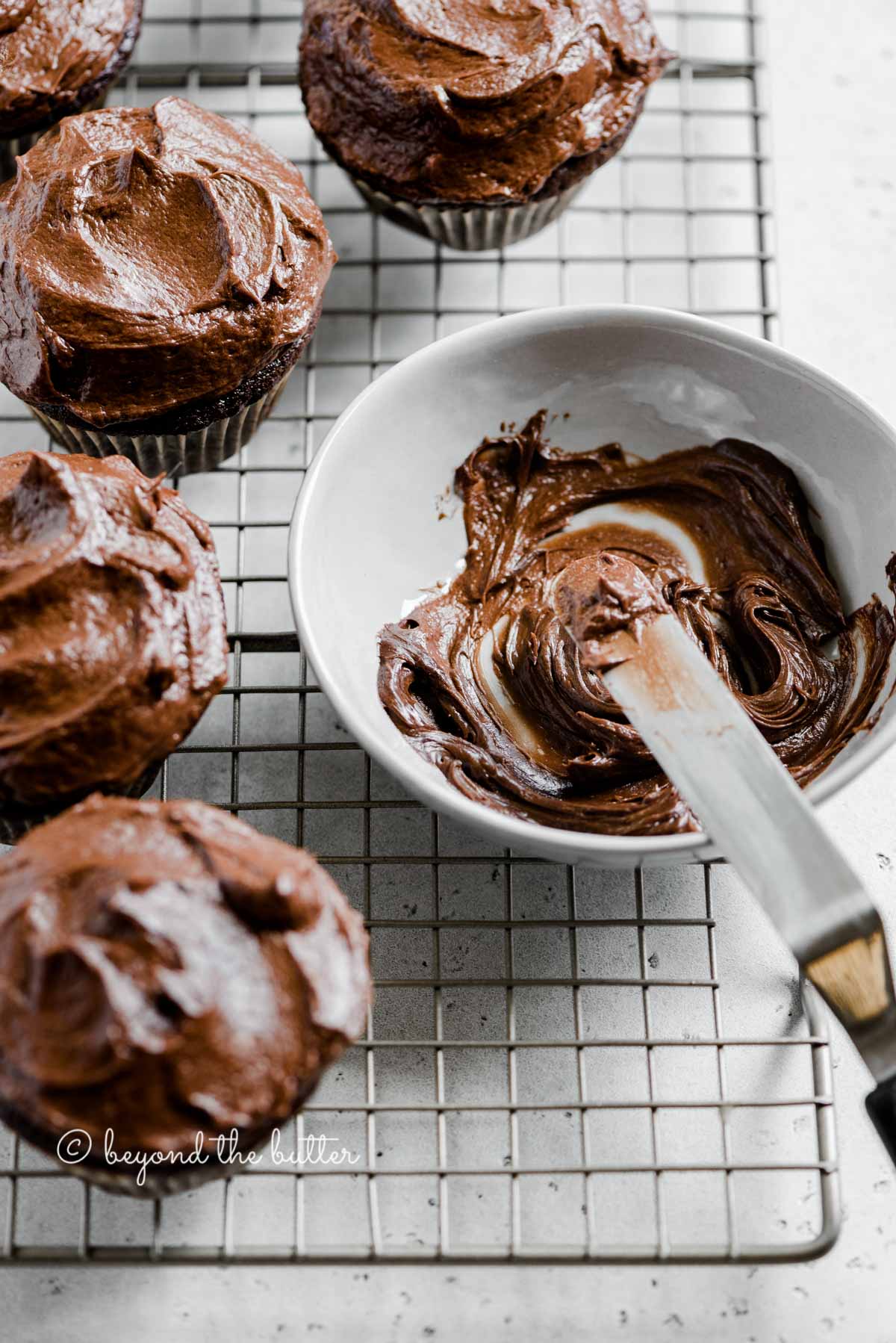 Angled image of chocolate cupcakes on cooling rack with small bowl of chocolate frosting and offset spatula | All Images © Luyiz™