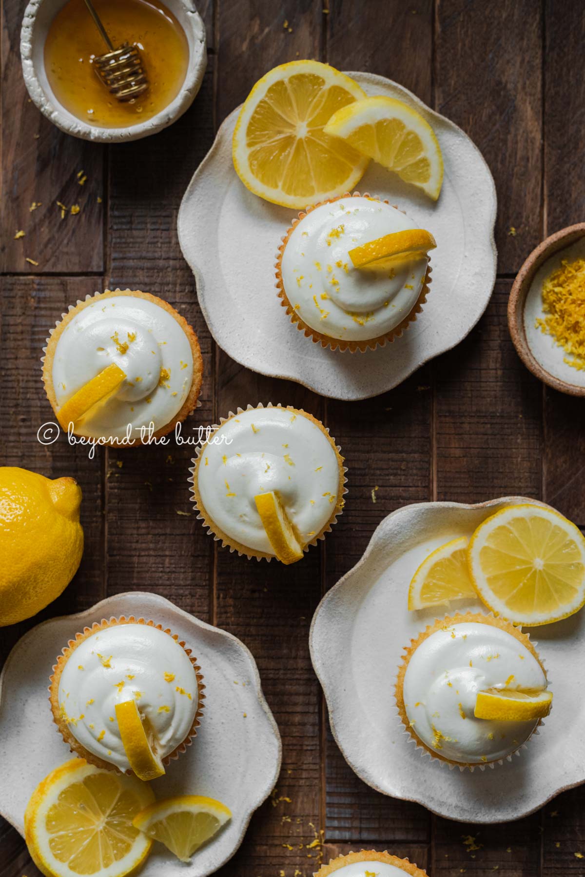 Randomly placed lemon cupcakes with lemon cream cheese frosting, garnished with lemon triangles on a dark wood background | All Images © Luyiz®