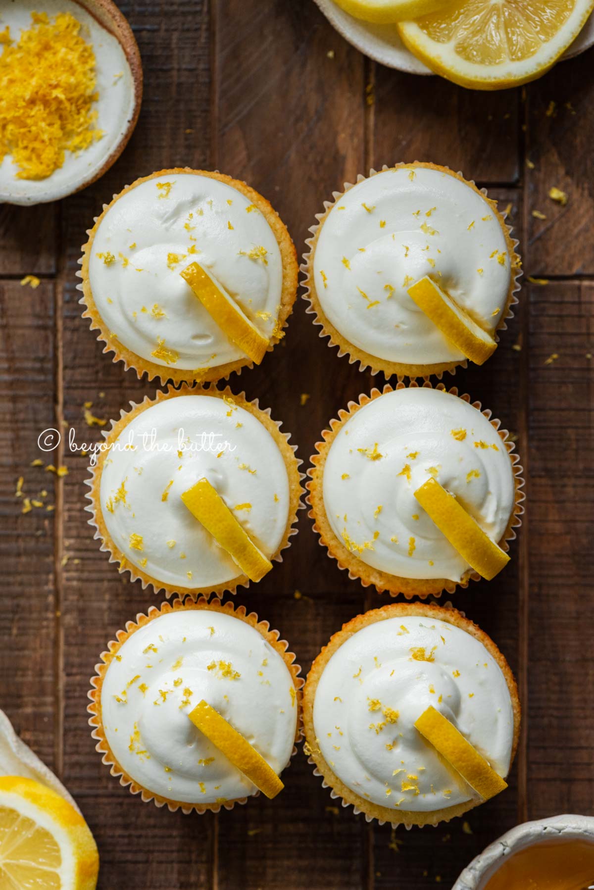 Two rows of small batch lemon cupcakes with lemon cream cheese frosting and garnished with lemon triangles on a dark wood background | All Images © Luyiz®