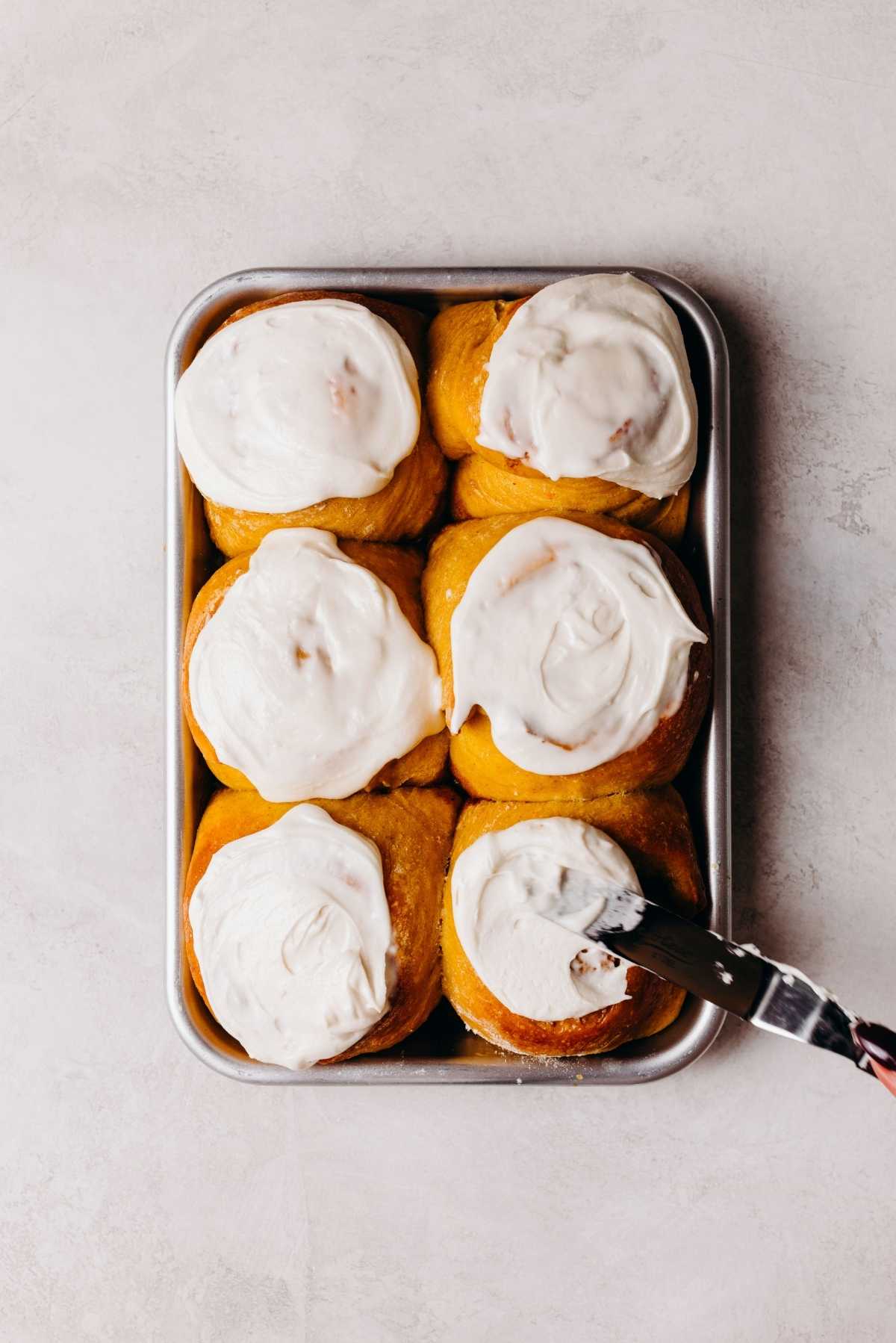 Pumpkin cinnamon rolls on a metal baking sheet being topped with maple frosting using an offset spatula.