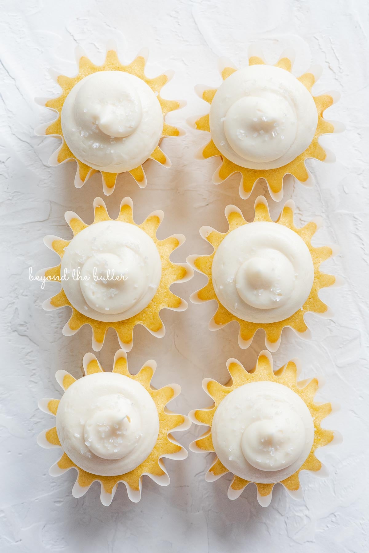 Overhead image of six small batch vanilla cupcakes with vanilla buttercream frosting sprinkled with sparkling sugar in two columns on white background.