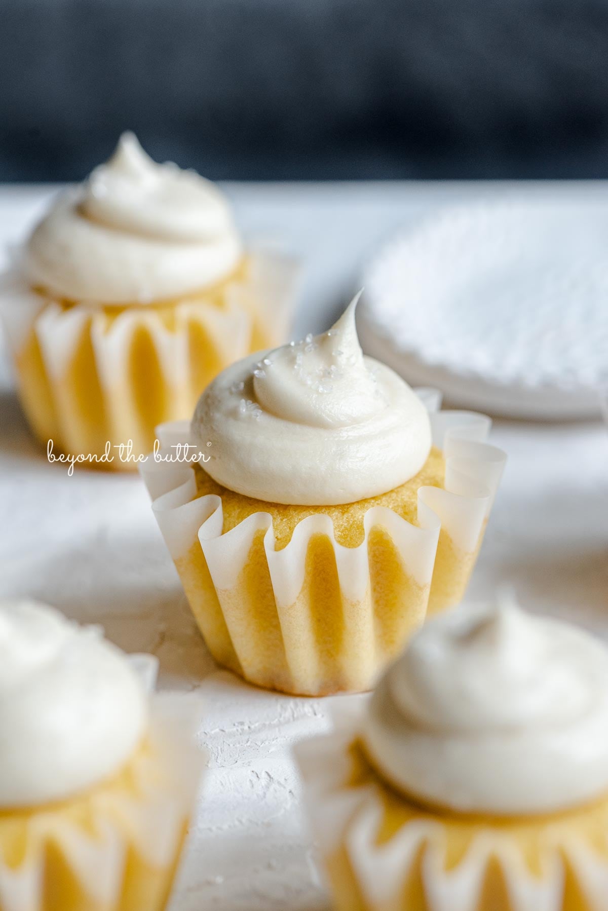 Angled image of frosted small batch vanilla cupcakes sprinkled with sparkling sugar randomly placed on white and dark blue background next to a small shallow white bowl of sparkling sugar.