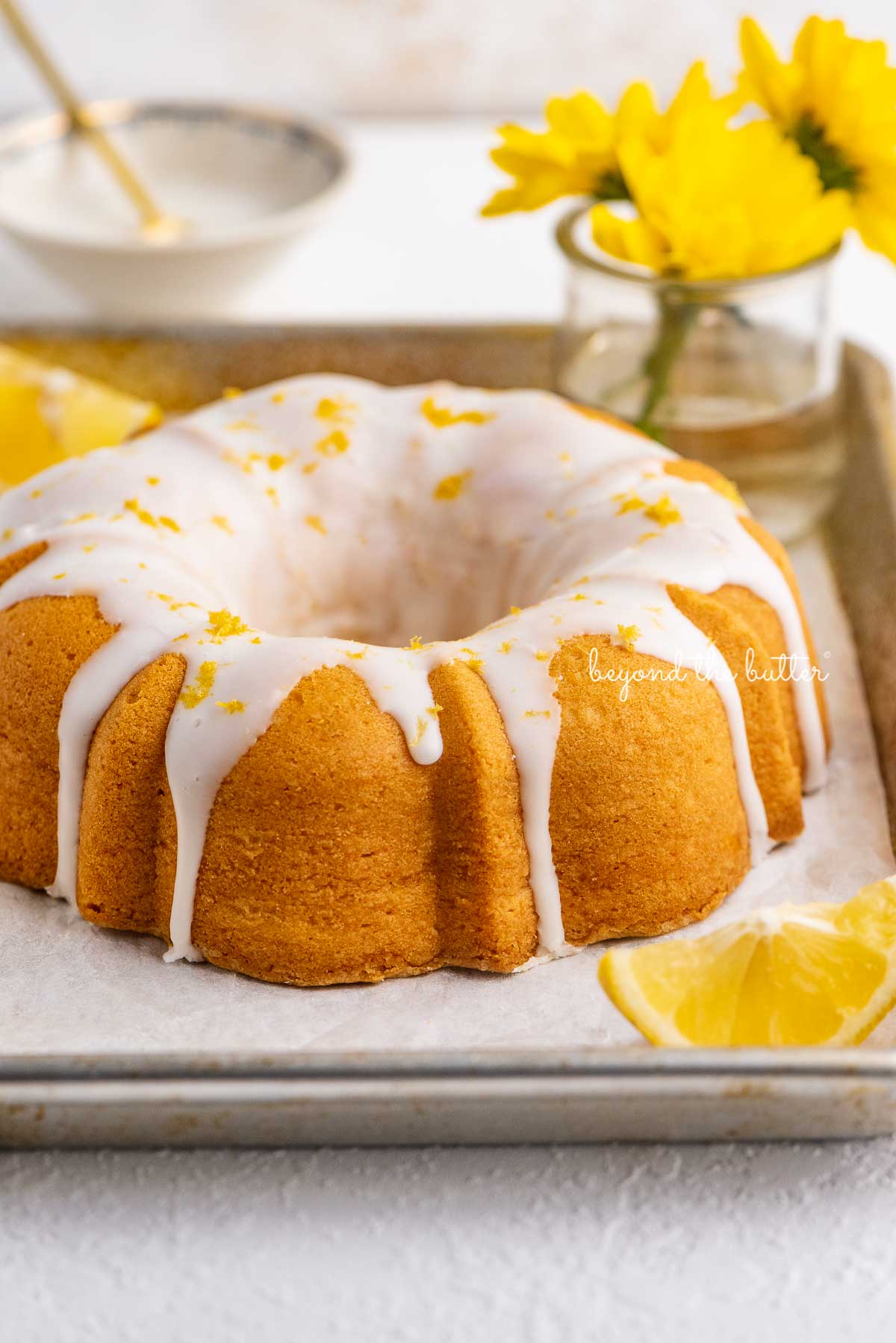 Small lemon cream cheese pound cake topped with a simple lemon glaze on a parchment paper lined baking sheet with sliced lemons and small bouquet of daisies | © Luyiz®
