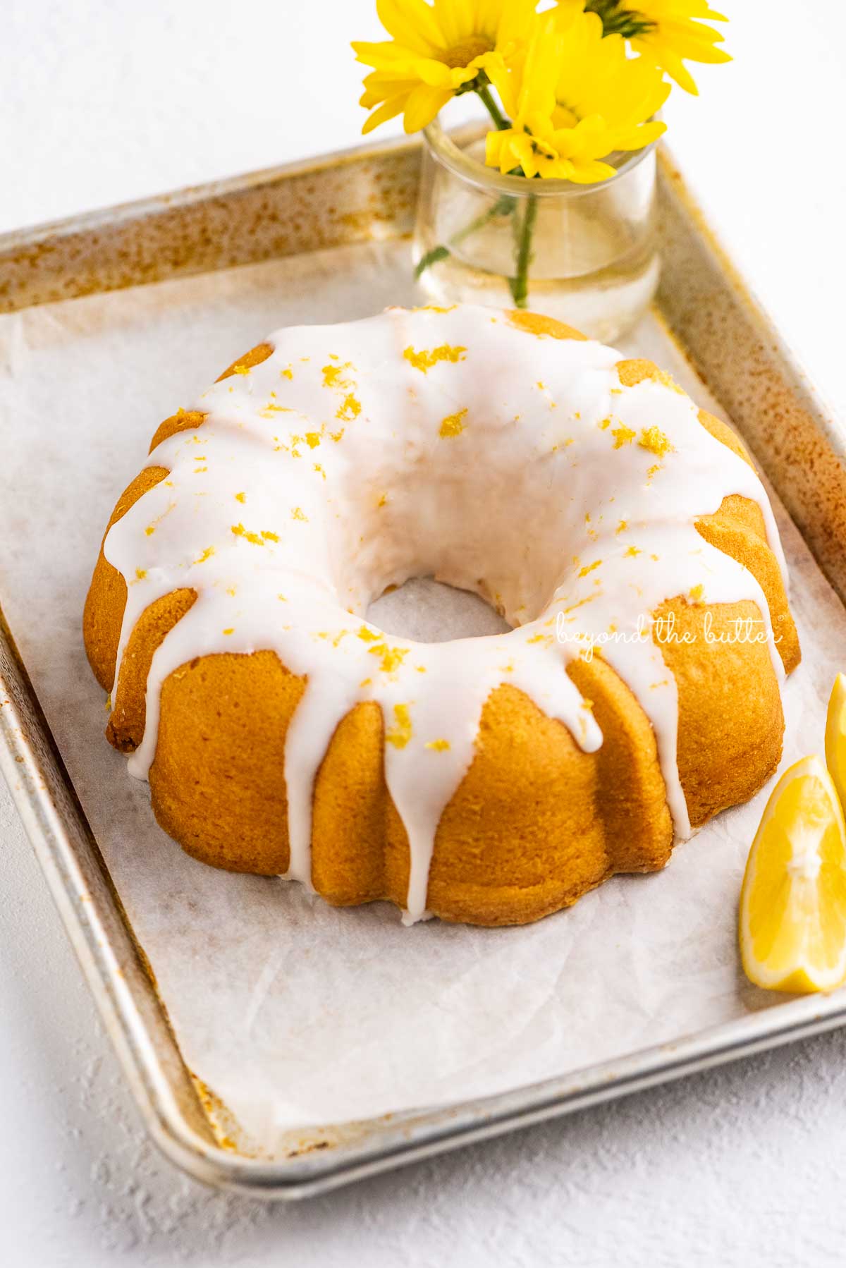 Small lemon cream cheese pound cake topped with a simple lemon glaze on a parchment paper lined baking sheet with sliced lemons and small bouquet of daisies | © Luyiz®