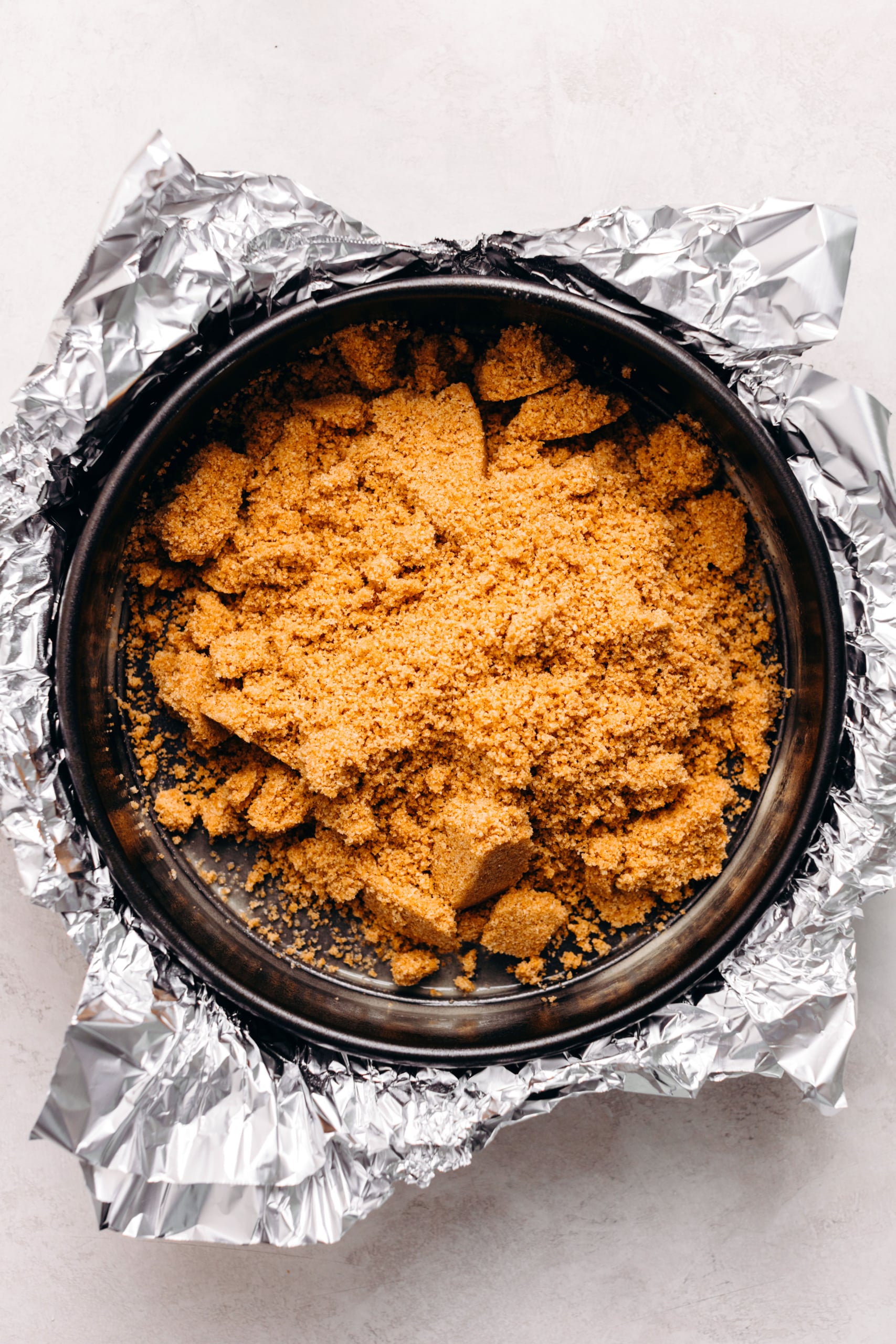 Graham cracker crust mixture placed on a aluminum foil wrapped springform pan on a textured white background.
