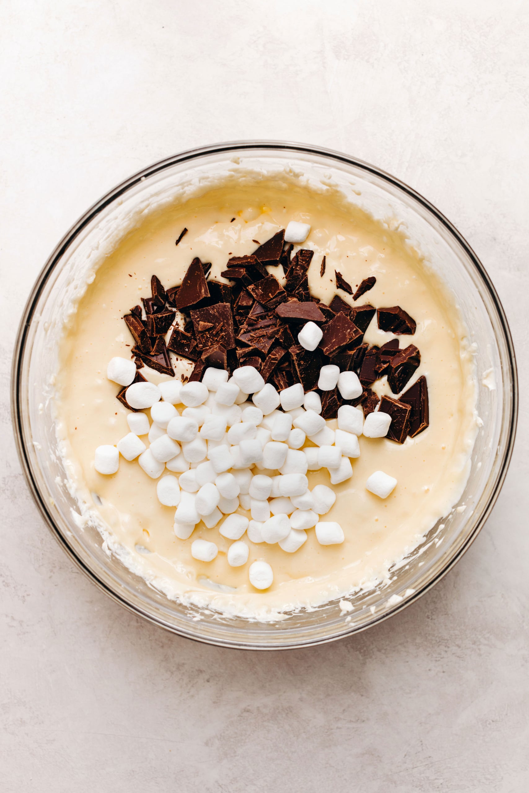Folding in mini marshmallows and chopped dark chocolate to cheesecaek batter in a large glass mixing bowl on a textured white background.