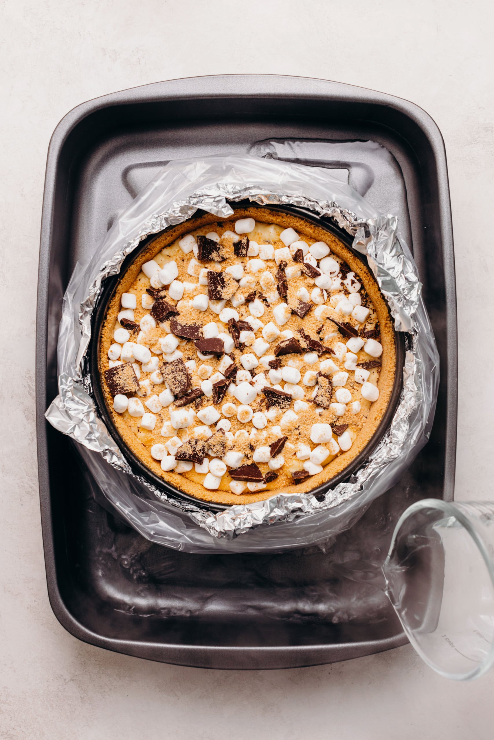 S'mores cheesecake topped with mini marshmallows, chopped dark chocolate, and graham cracker crumbs wrapped in aluminum foil and a slow cooker liner bag placed in a roasting pan with boiling hot water.