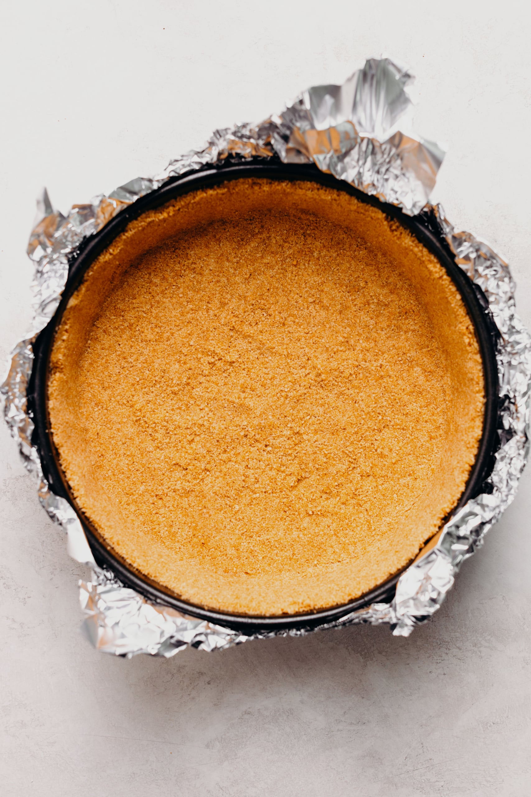 Baked graham cracker crust in a aluminum foil wrapped springform pan on a textured white background.