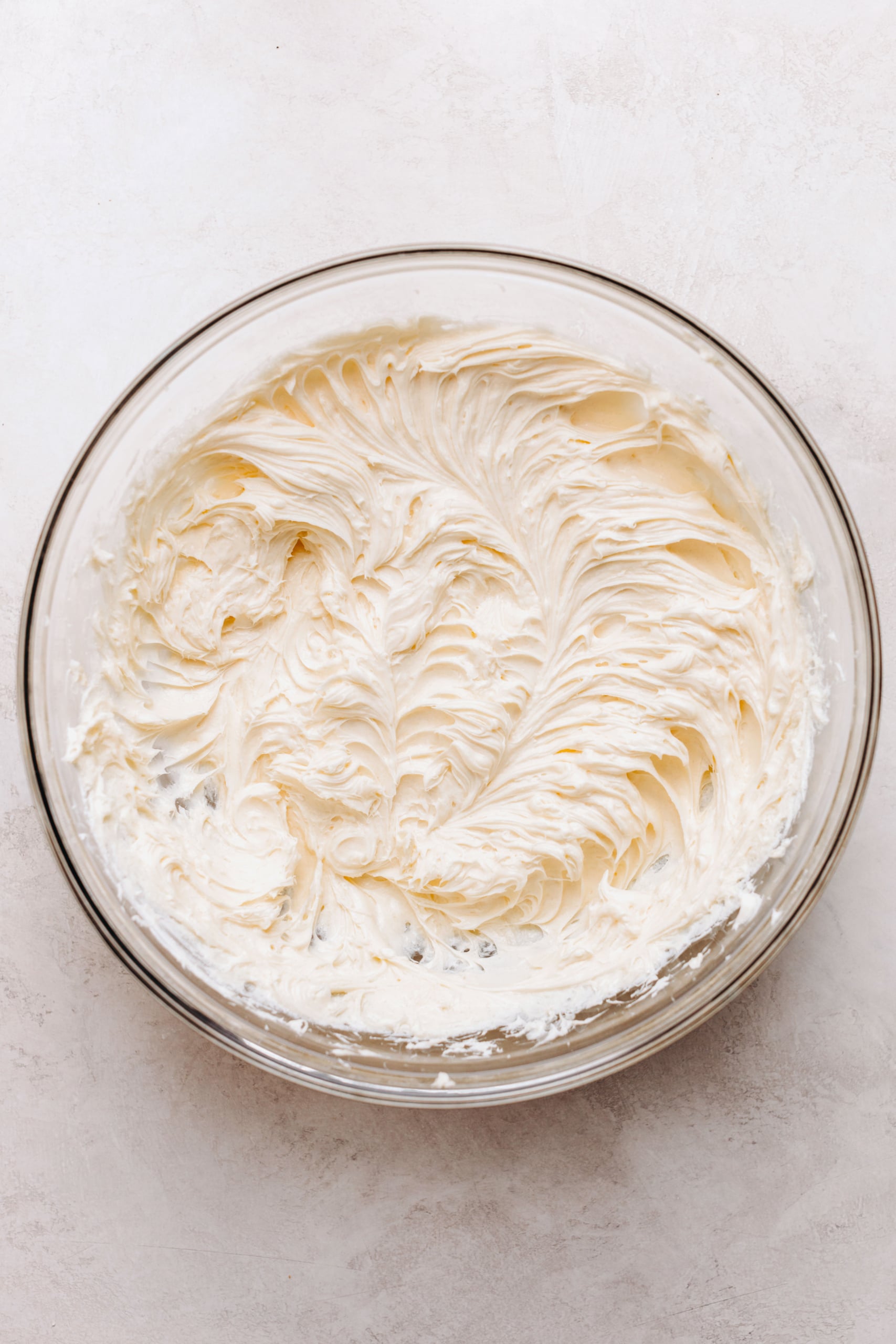 Cream cheese, granulated sugar, and vanilla extract blended together in a large glass mixing bowl on a textured white background.