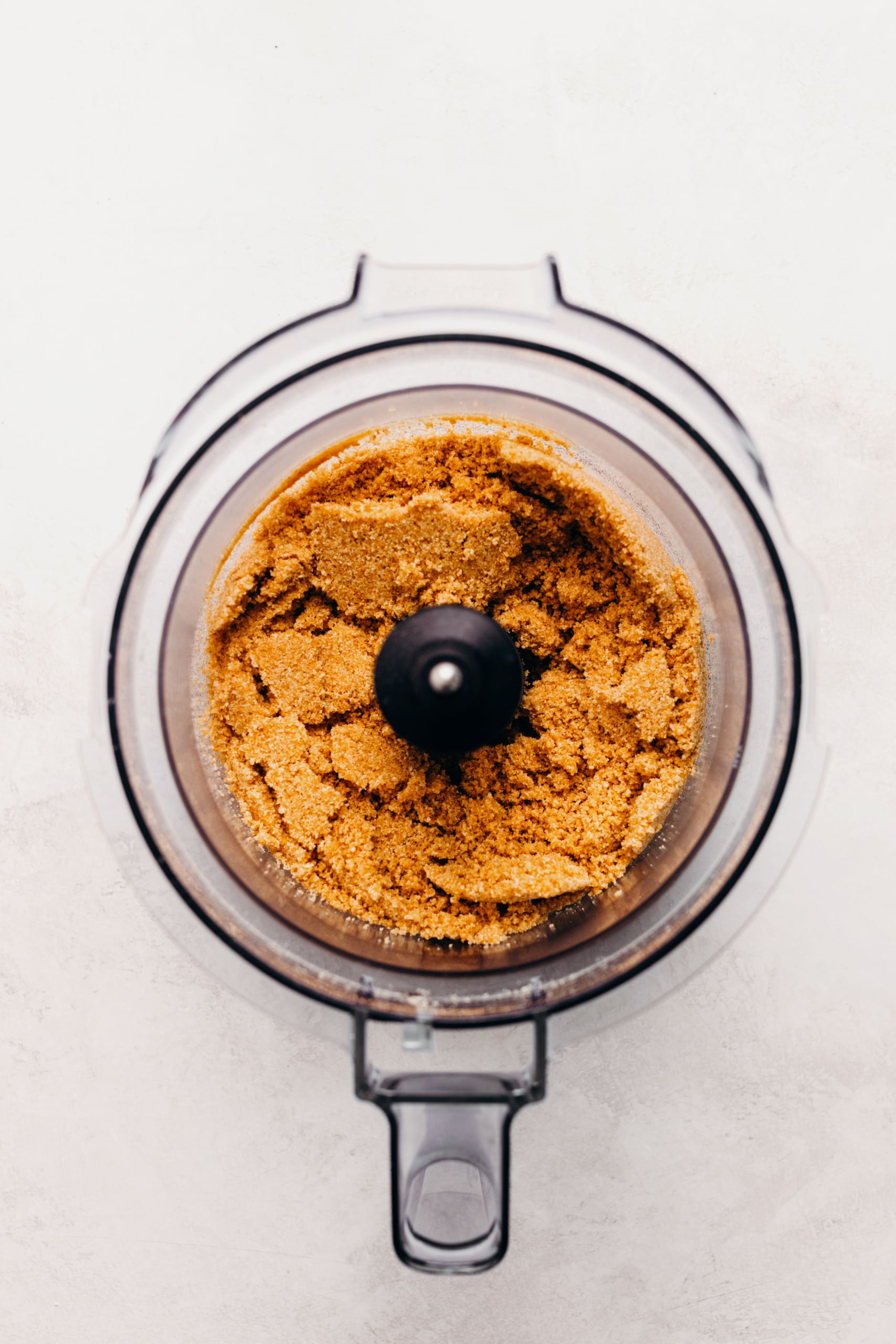 Graham cracker crumbs mixed together with melted butter and sugar in a food processor on a textured white background.