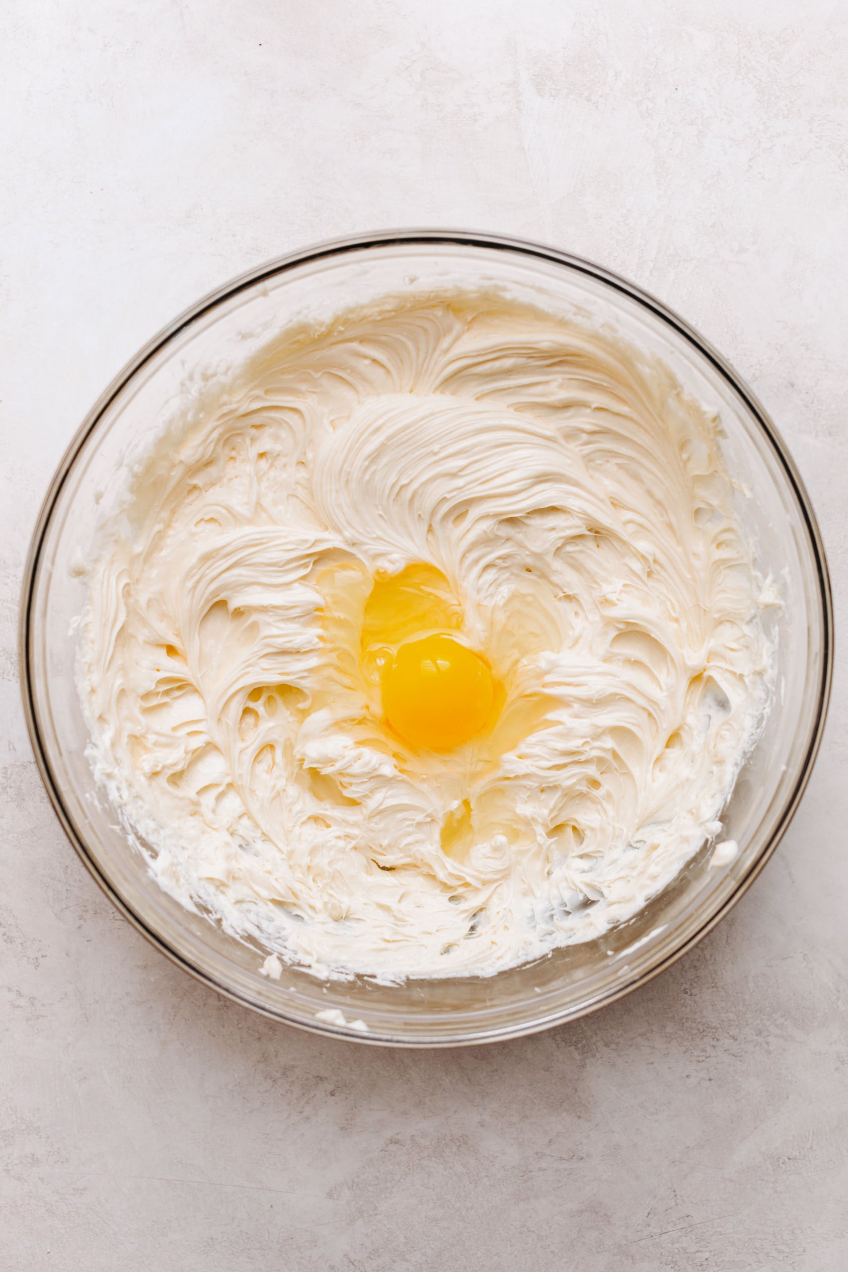 Adding eggs to cheesecake batter in a large glass mixing bowl on a textured white background.