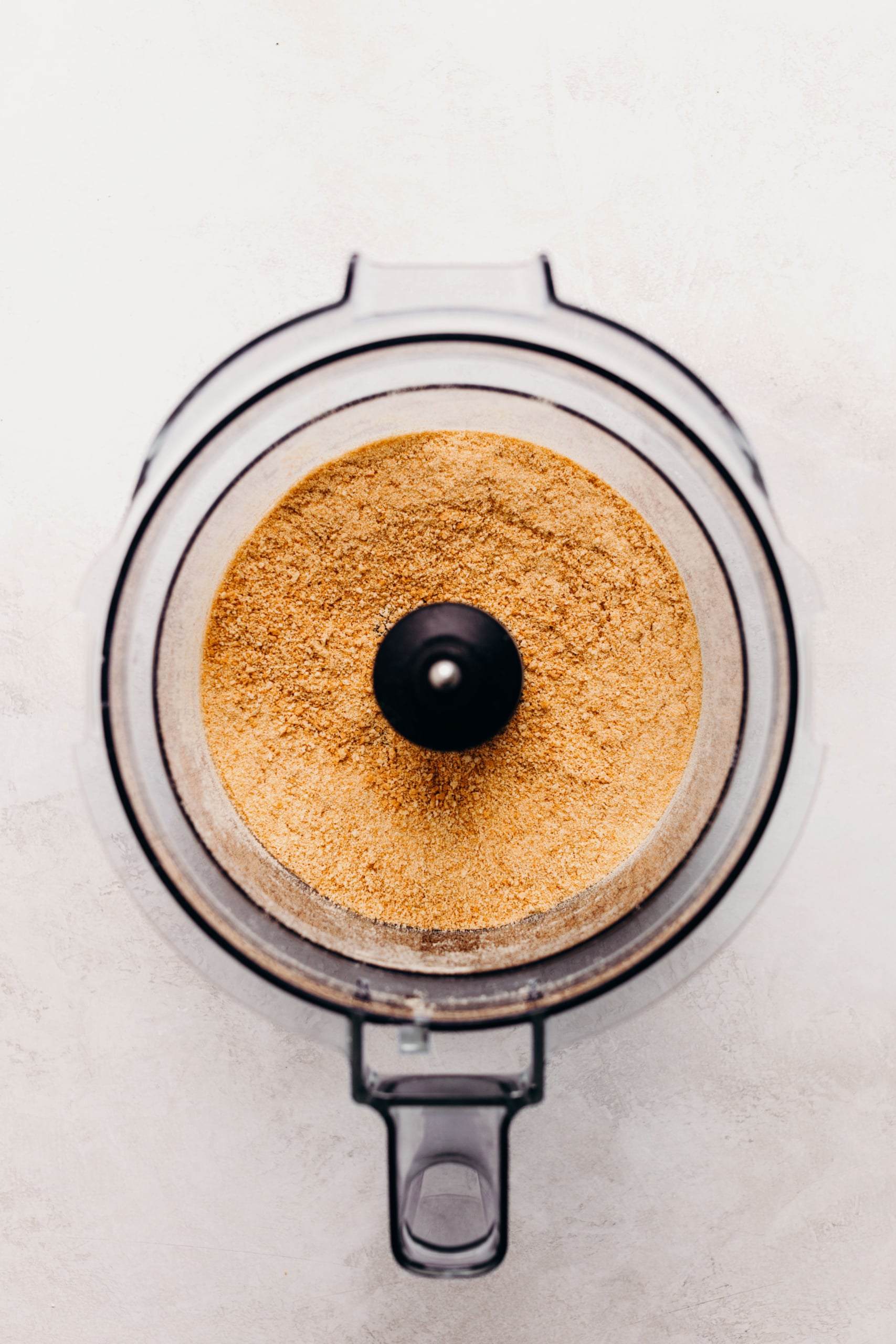 Graham cracker crumbs in a food processor on a textured white background.
