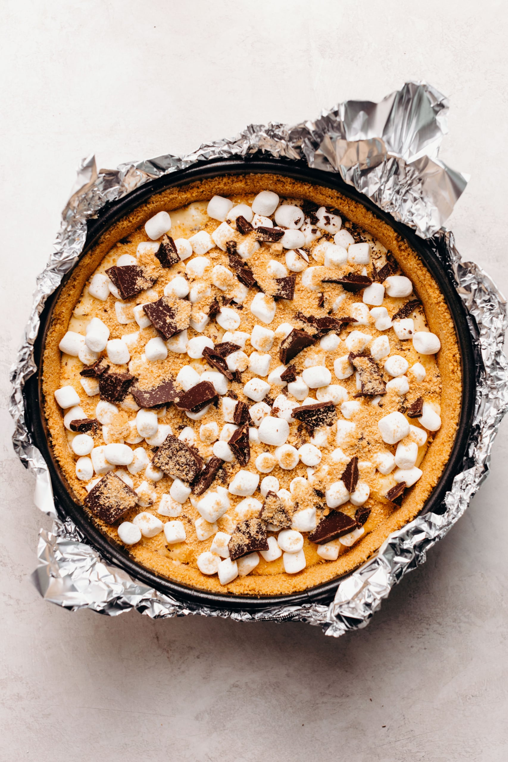 Unbaked s'mores cheesecake topped with mini marshmallows, chopped dark chocolate, and graham cracker crumbs in an aluminum foil wrapped springform pan on a white textured background.
