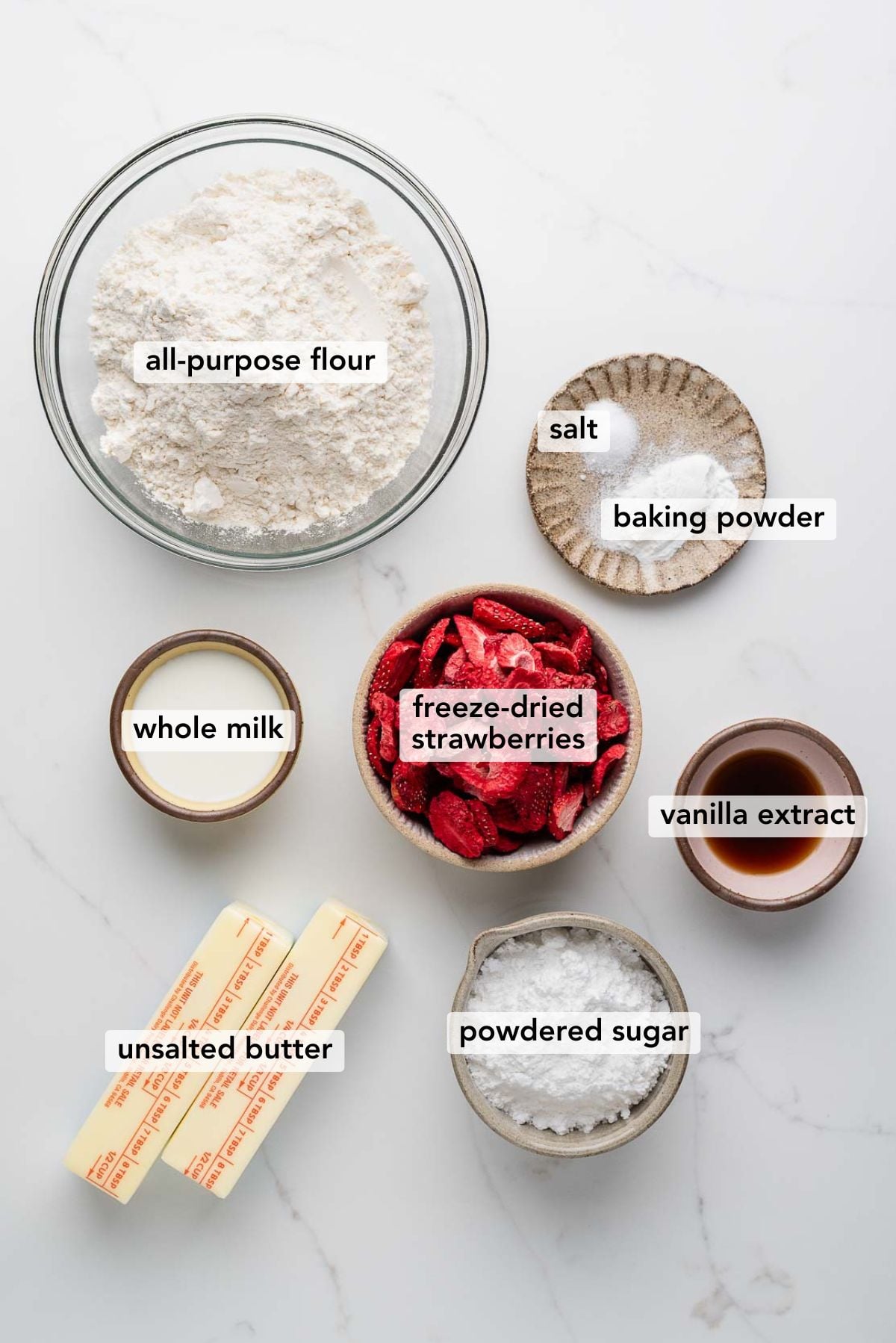 Ingredients in various size bowls and plates labeled as all purpose flour, salt, baking powder, whole milk, freeze dried strawberries, vanilla extract, unsalted butter, and powdered sugar on a white marbled background.