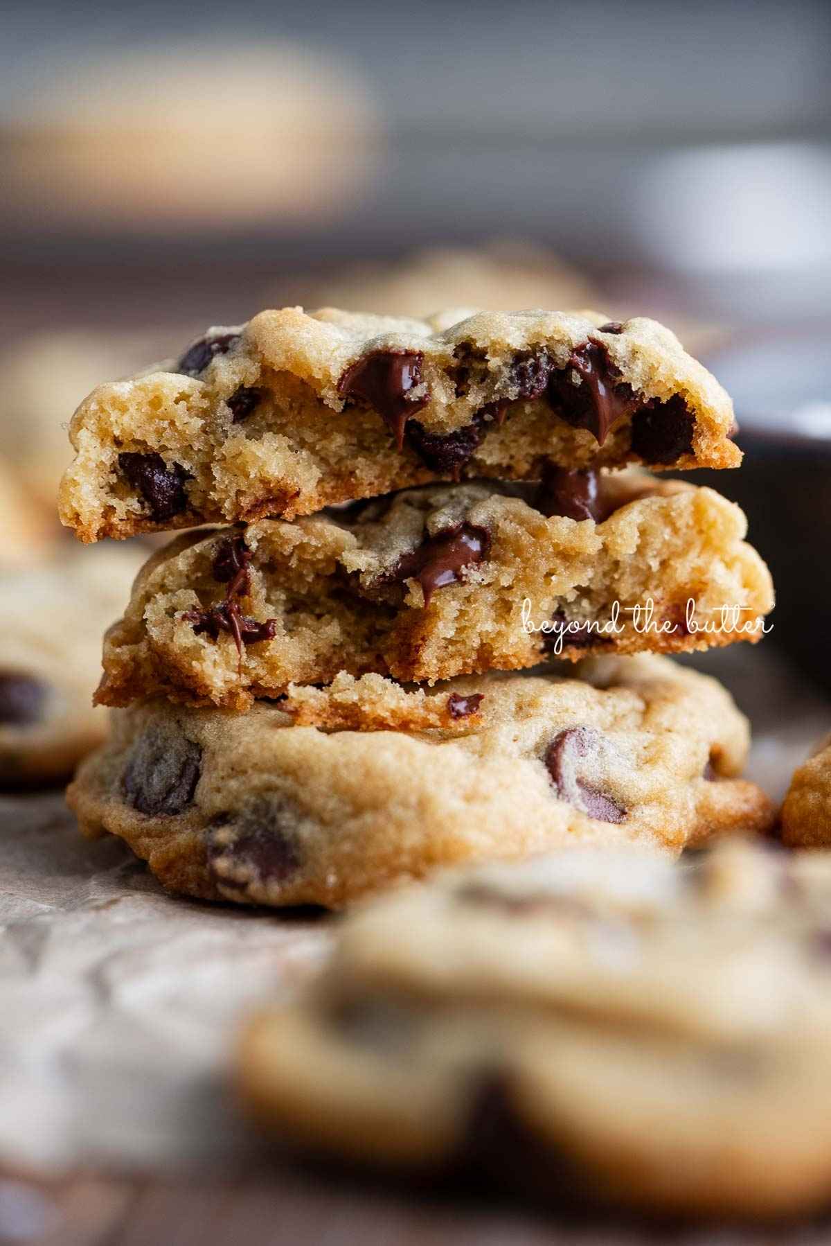 Chocolate chip cookies randomly placed on crumpled parchment paper with two cookies stacked on top of the other. The top cookie is split in half to see the gooey center.