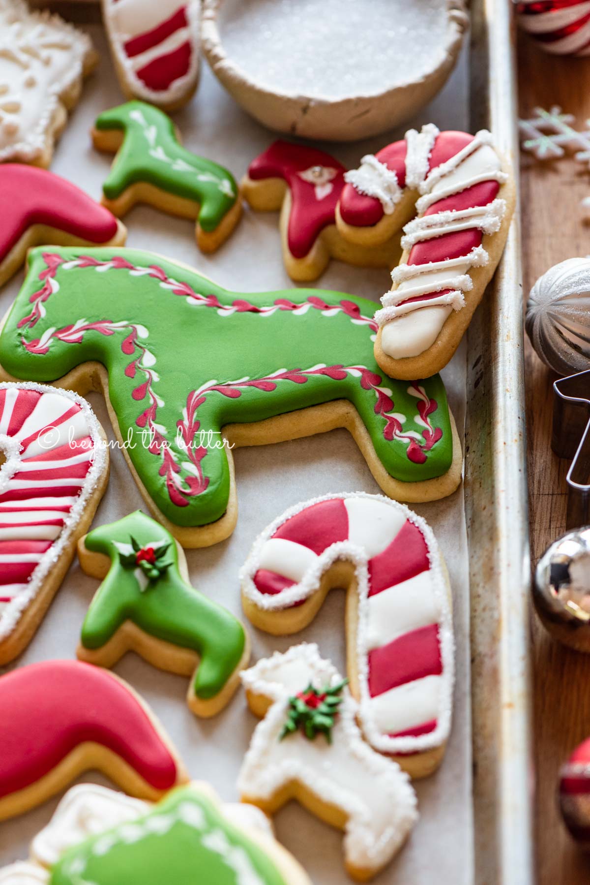 Randomly placed decorated cut out sugar cookies on a parchment lined baking sheet with ornaments and cookie cutters around it | All Images © Luyiz®