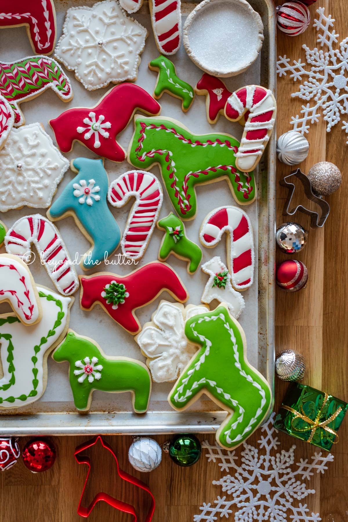 Randomly placed decorated cut out sugar cookies on a parchment lined baking sheet with ornaments and cookie cutters around it | All Images © Luyiz®