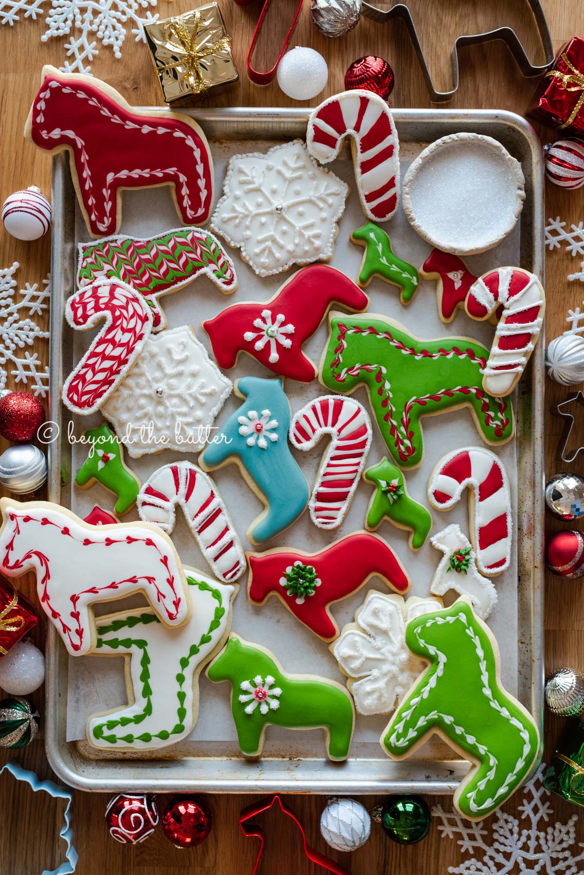 Randomly placed decorated cut out sugar cookies on a parchment lined baking sheet with ornaments and cookie cutters around it | All Images © Luyiz®