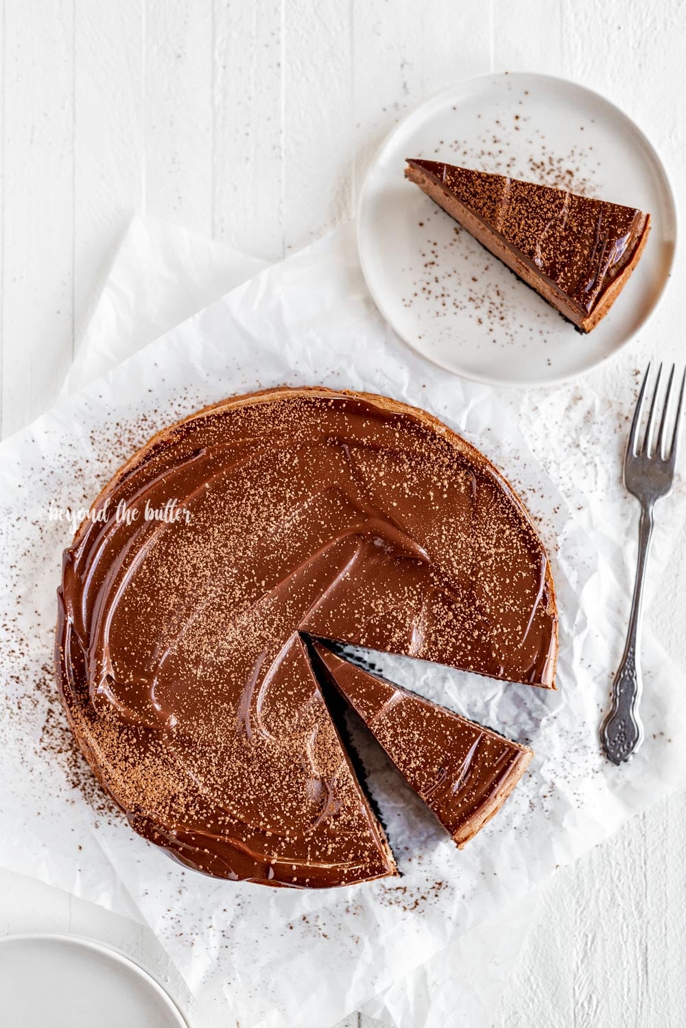 Overhead image of Triple Chocolate Mocha Cheesecake dusted with cocoa and a slice removed set on a dessert plate with a fork | All Images © Luyiz, LLC