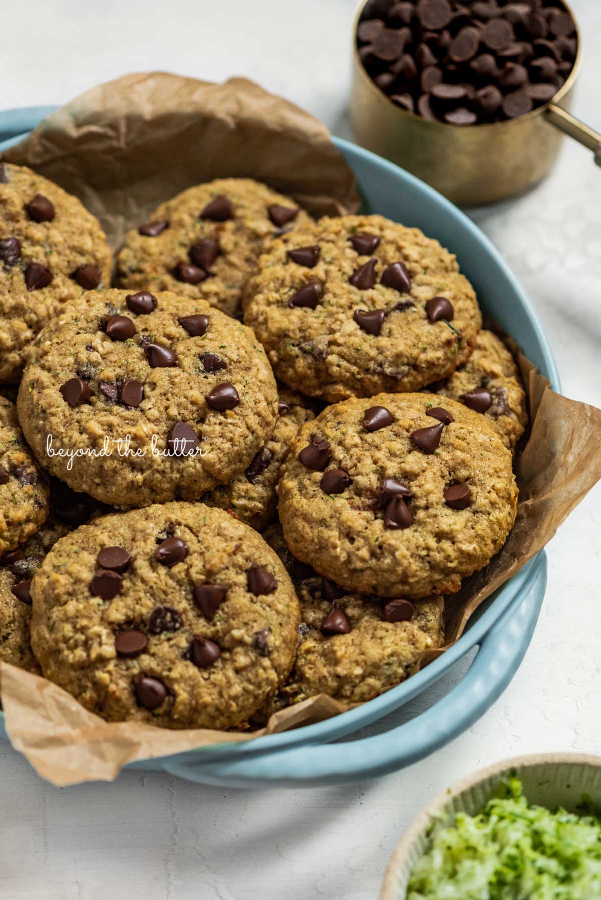 Baked zucchini chocolate chip oatmeal cookies in a parchment paper lined blue serving dish with a measuring cup of chocolate chips and a bowl of shredded zucchini nearby | © Luyiz®