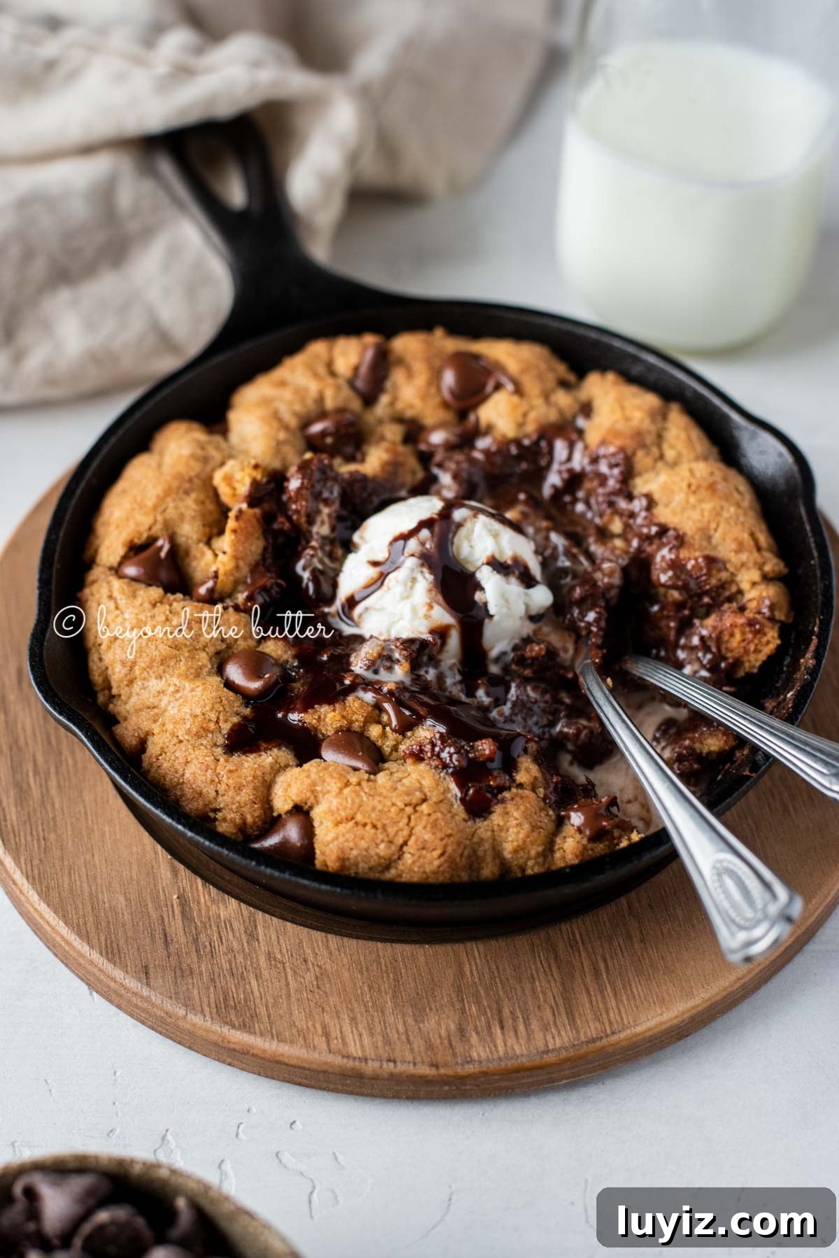 Brown Butter Chocolate Chip Skillet Cookie Recipe 2 Warm chocolate chip skillet cookie with scoop of vanilla ice cream and two spoons resting in skillet on wood trivet
