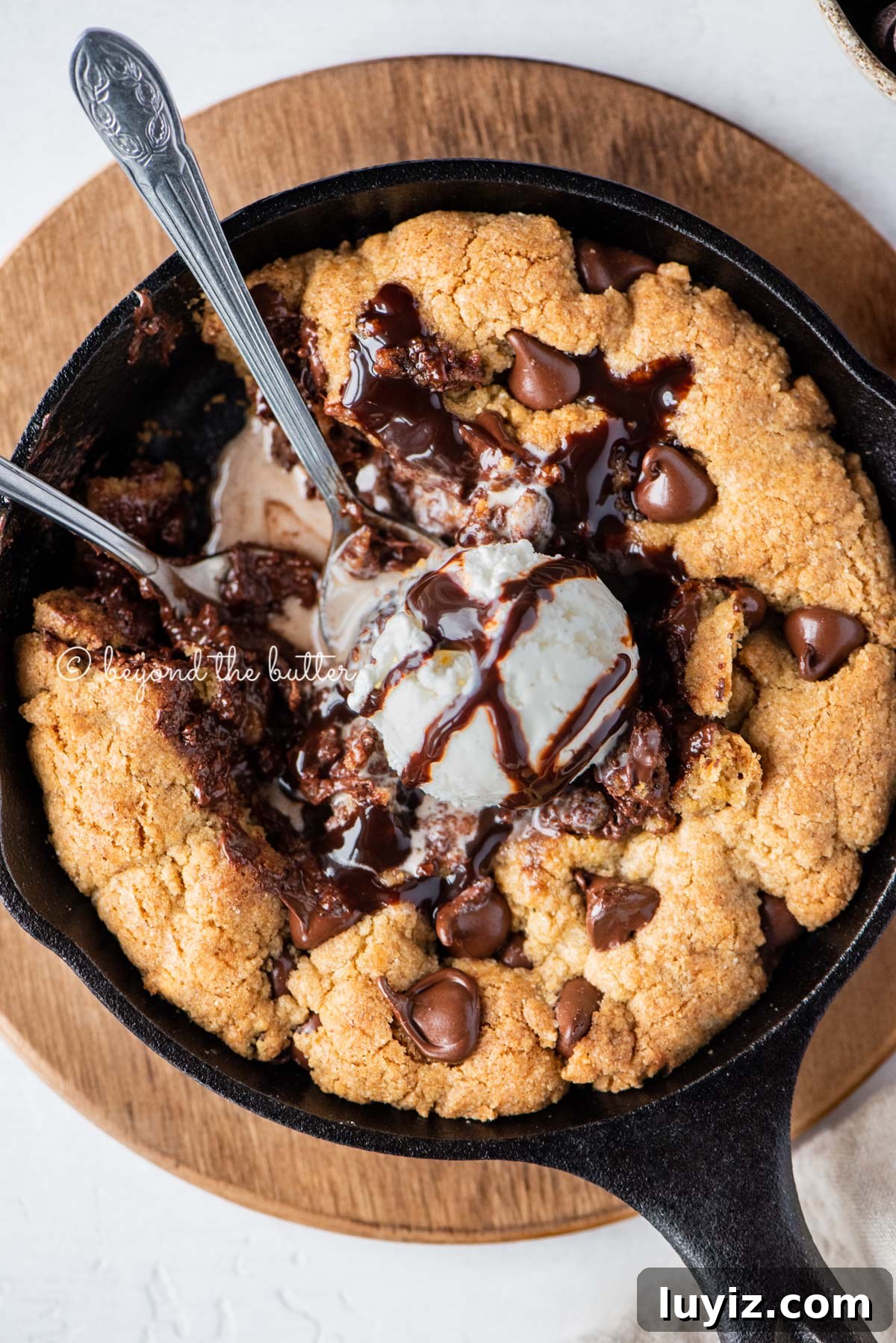 Brown Butter Chocolate Chip Skillet Cookie Recipe 4 Closeup of brown butter chocolate chip skillet cookie with scoop of vanilla ice cream and two spoons resting in skillet on wood trivet