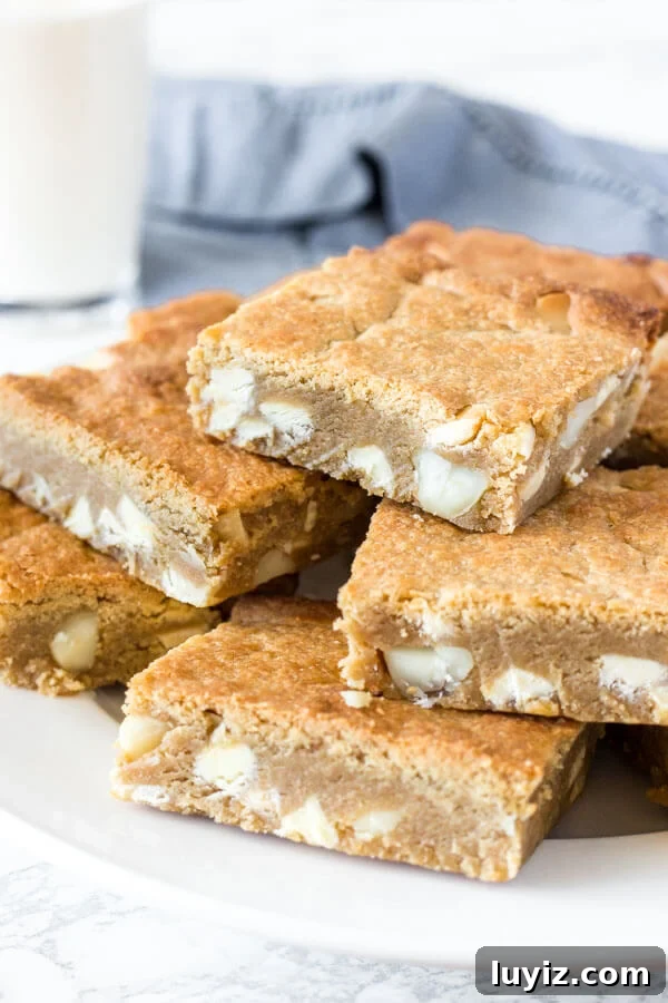 White Chocolate Macadamia Blondie Recipe — Chewy & Decadent 3 A plate of fudgy white chocolate macadamia nut blondies with a glass of milk in the background.