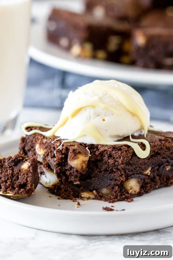 White Chocolate Macadamia Nut Brownies Recipe 5 A fudgy white chocolate macadamia brownie with a scoop of vanilla ice cream and melted white chocolate drizzle with a plate of brownies in the background.