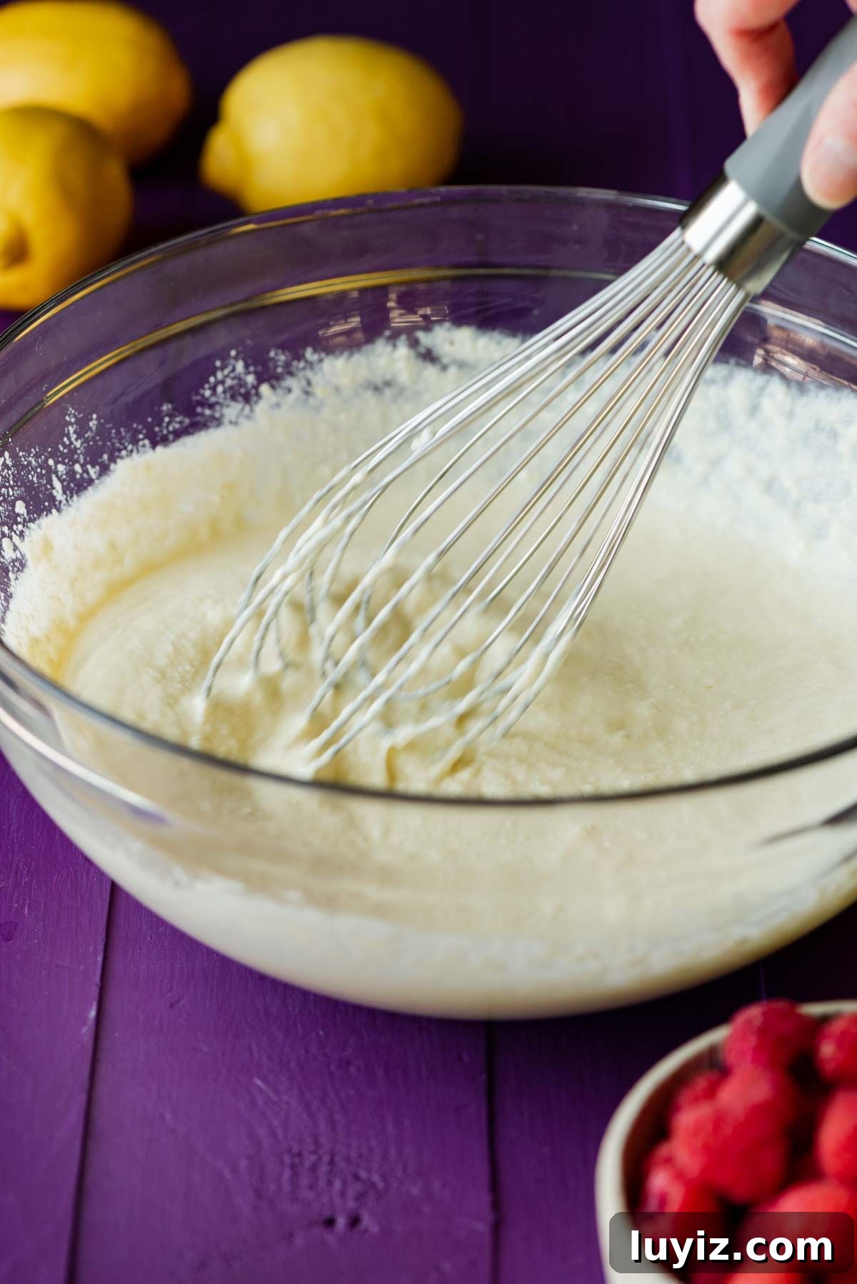 Lemon Raspberry Ricotta Cake Recipe — Moist Spring Dessert 6 Whisking the wet ingredients in a glass bowl to make lemon raspberry ricotta cake on a purple background.