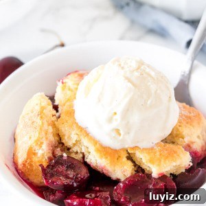 A bowl of fresh cherry cobbler with a flaky biscuit topping and scoop of vanilla ice cream on top.