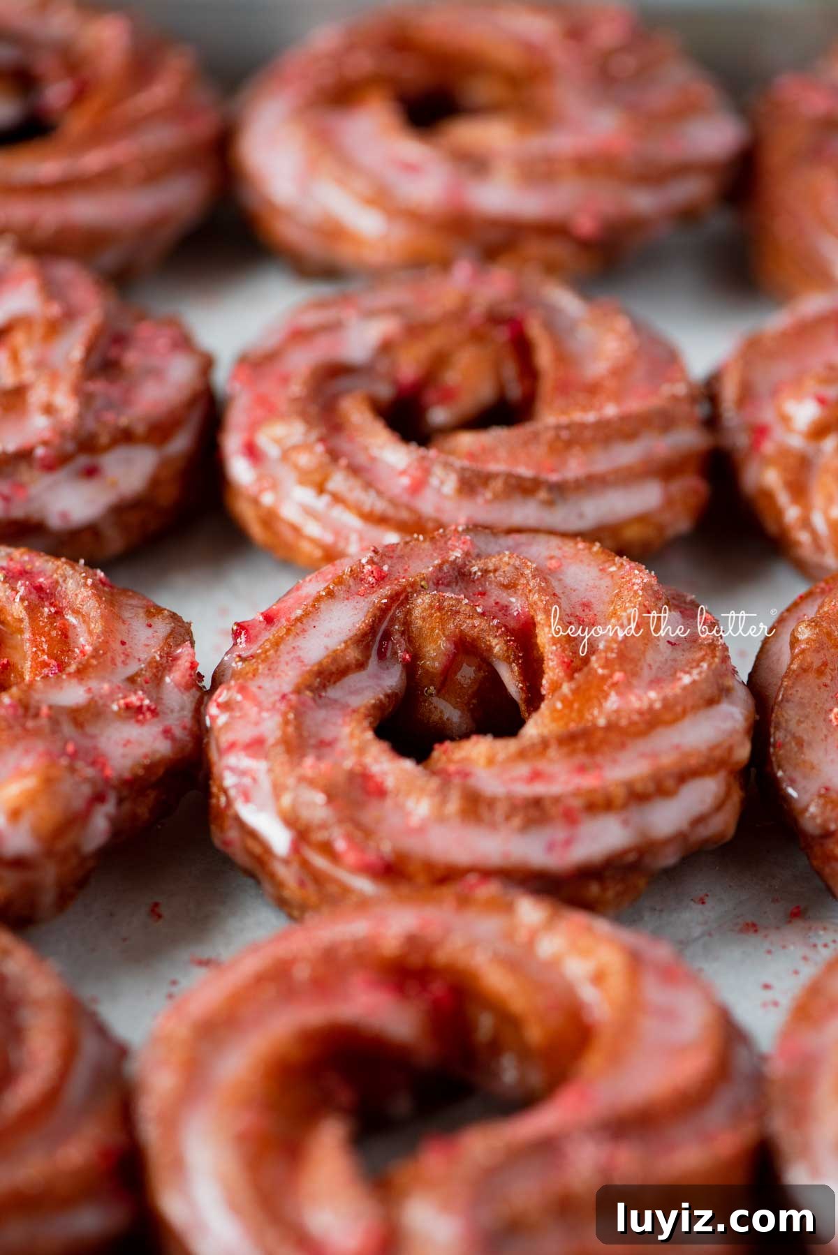 Strawberry-Glazed French Crullers Recipe for Homemade Pastries 6 Strawberry glazed french crullers sprinkled with strawberry sugar on a parchment paper lined baking sheet | © Beyond the Butter®