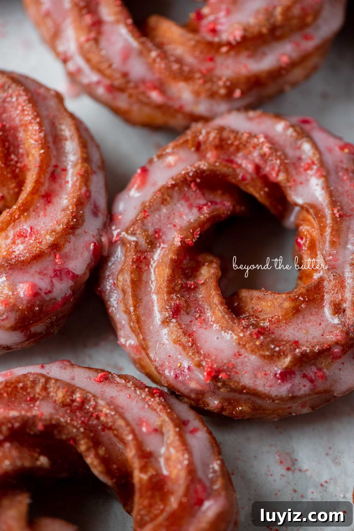 Strawberry-Glazed French Crullers Recipe for Homemade Pastries 8 Strawberry glazed french crullers sprinkled with strawberry sugar on a parchment paper lined baking sheet | © Beyond the Butter®