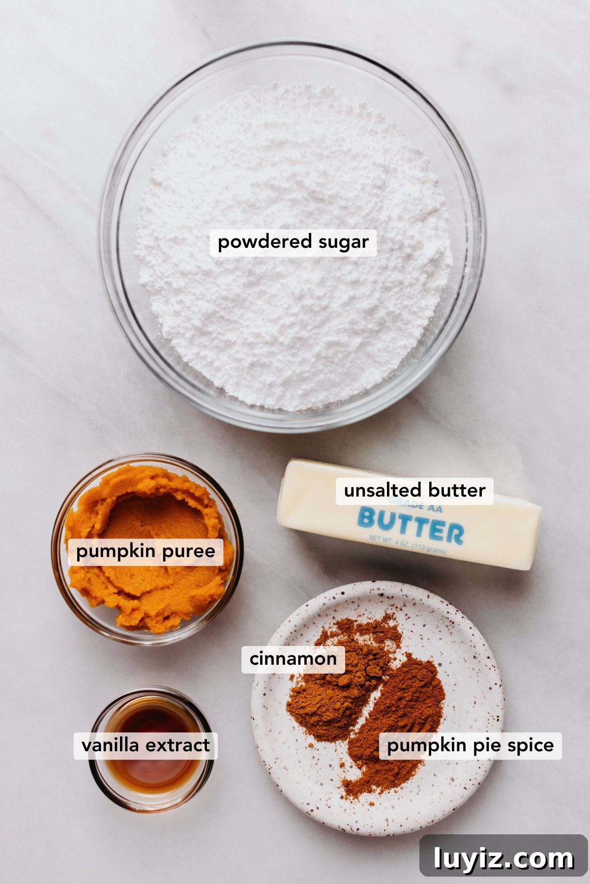 Pumpkin Fudge Brownies with Creamy Frosting 5 Ingredients in various size bowls labeled as powdered sugar, pumpkin puree, unsalted butter, vanilla extract, cinnamon, and pumpkin pie spice on a white marbled background.