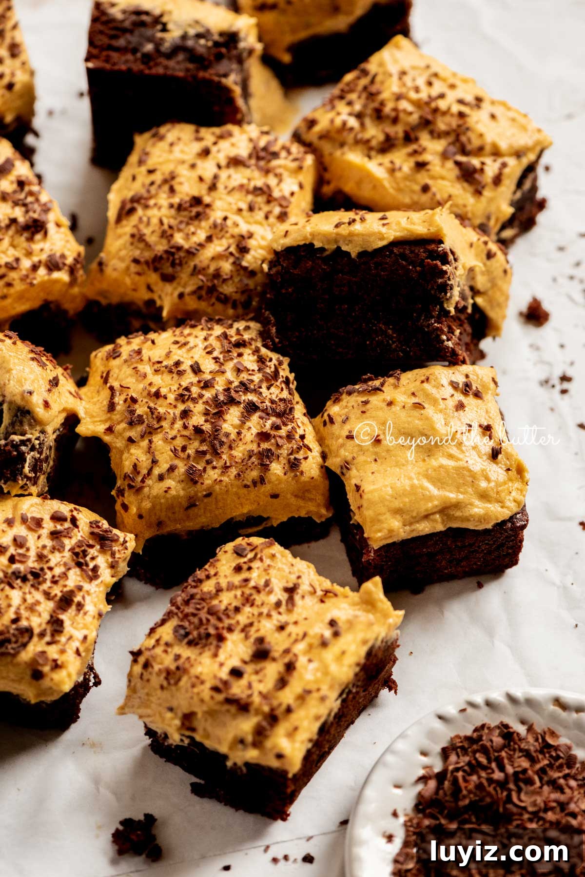 Pumpkin Fudge Brownies with Creamy Frosting 6 pumpkin frosted brownies randomly placed on parchment paper with a side bowl of chocolate shavings