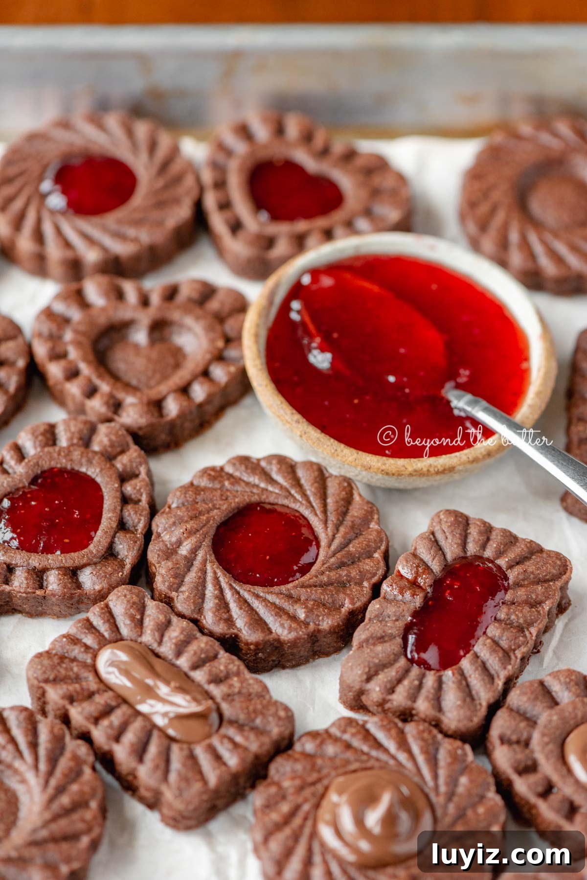 Chocolate Thumbprint Cookies with Raspberry Jam Filling 3 Parchment lined baking sheet full of chocolate thumbprint cookies with small bowl of strawberry jam and spoon | All Images © Beyond the Butter®