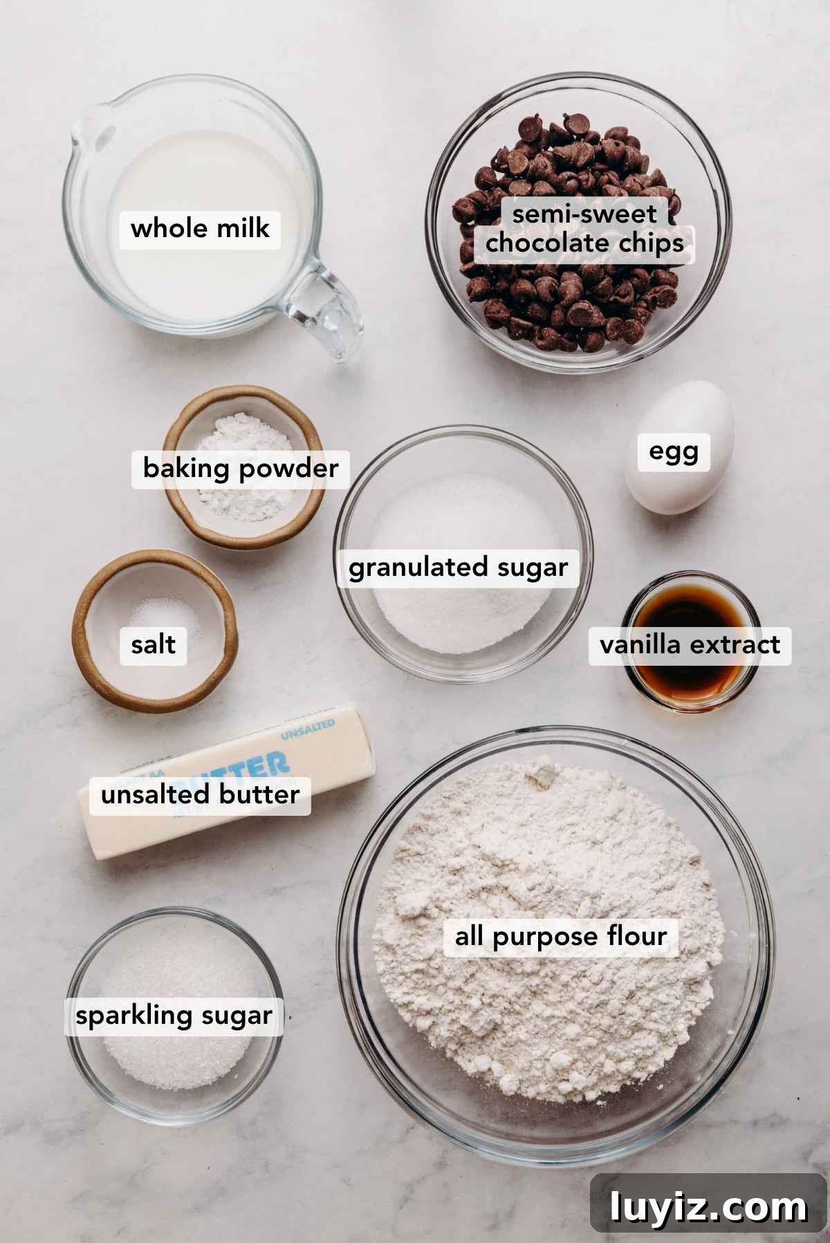 Crispy Chocolate Chip Scones with Vanilla Glaze 4 Ingredients in various size bowls labeled as whole milk, semi sweet chocolate chips, baking powder, granulated sugar, egg, salt, vanilla extract, unsalted butter, sparkling sugar, and all purpose flour on a white marbled background.