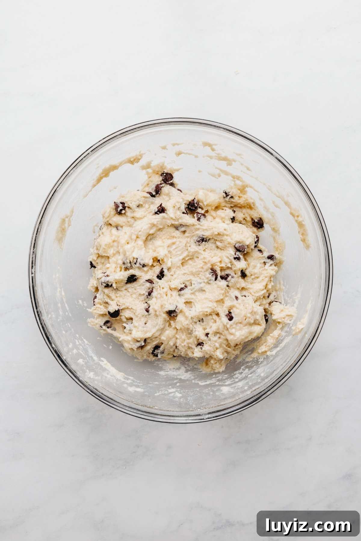 Crispy Chocolate Chip Scones with Vanilla Glaze 8 Chocolate chips mixed into scones mixture in a large glass mixing bowl on a white and grey marbled background.