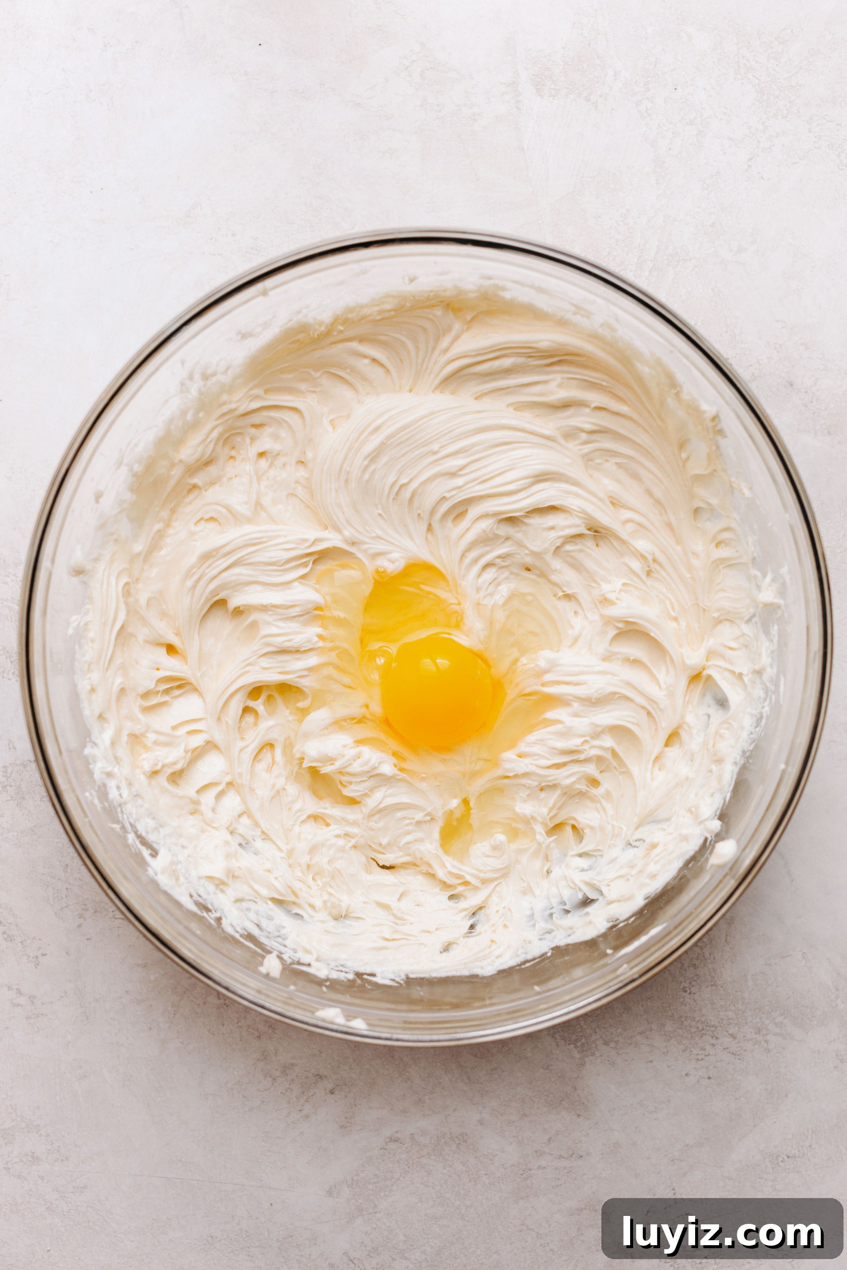 S’mores Cheesecake Recipe: Gooey Chocolate, Graham & Toasted Marshmallow 11 Adding eggs to cheesecake batter in a large glass mixing bowl on a textured white background.