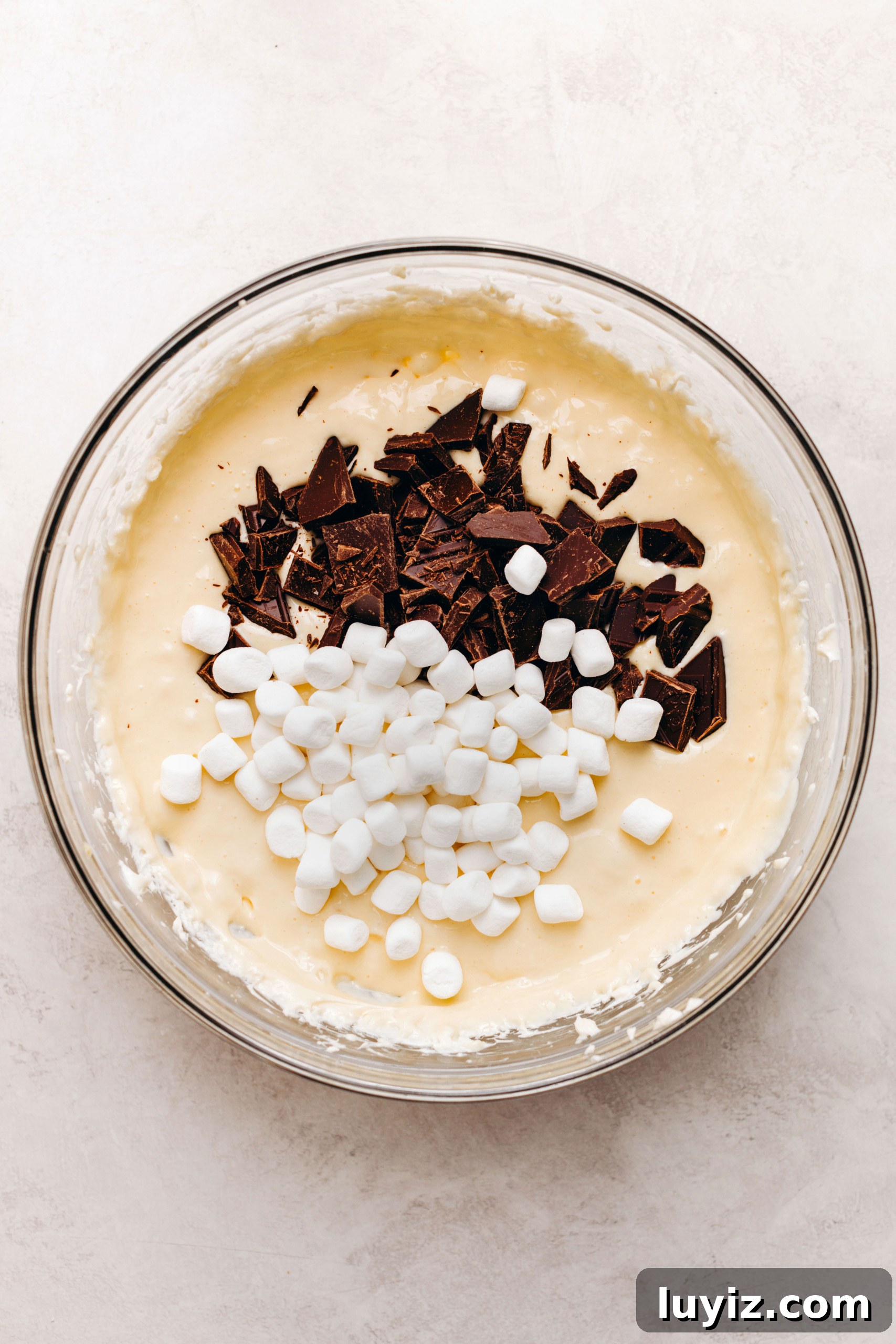 S’mores Cheesecake Recipe: Gooey Chocolate, Graham & Toasted Marshmallow 12 Folding in mini marshmallows and chopped dark chocolate to cheesecaek batter in a large glass mixing bowl on a textured white background.