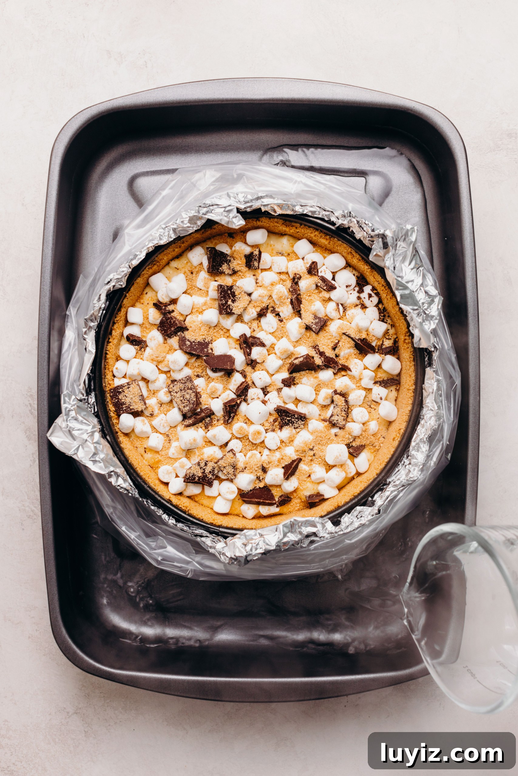S’mores Cheesecake Recipe: Gooey Chocolate, Graham & Toasted Marshmallow 14 S'mores cheesecake topped with mini marshmallows, chopped dark chocolate, and graham cracker crumbs wrapped in aluminum foil and a slow cooker liner bag placed in a roasting pan with boiling hot water.