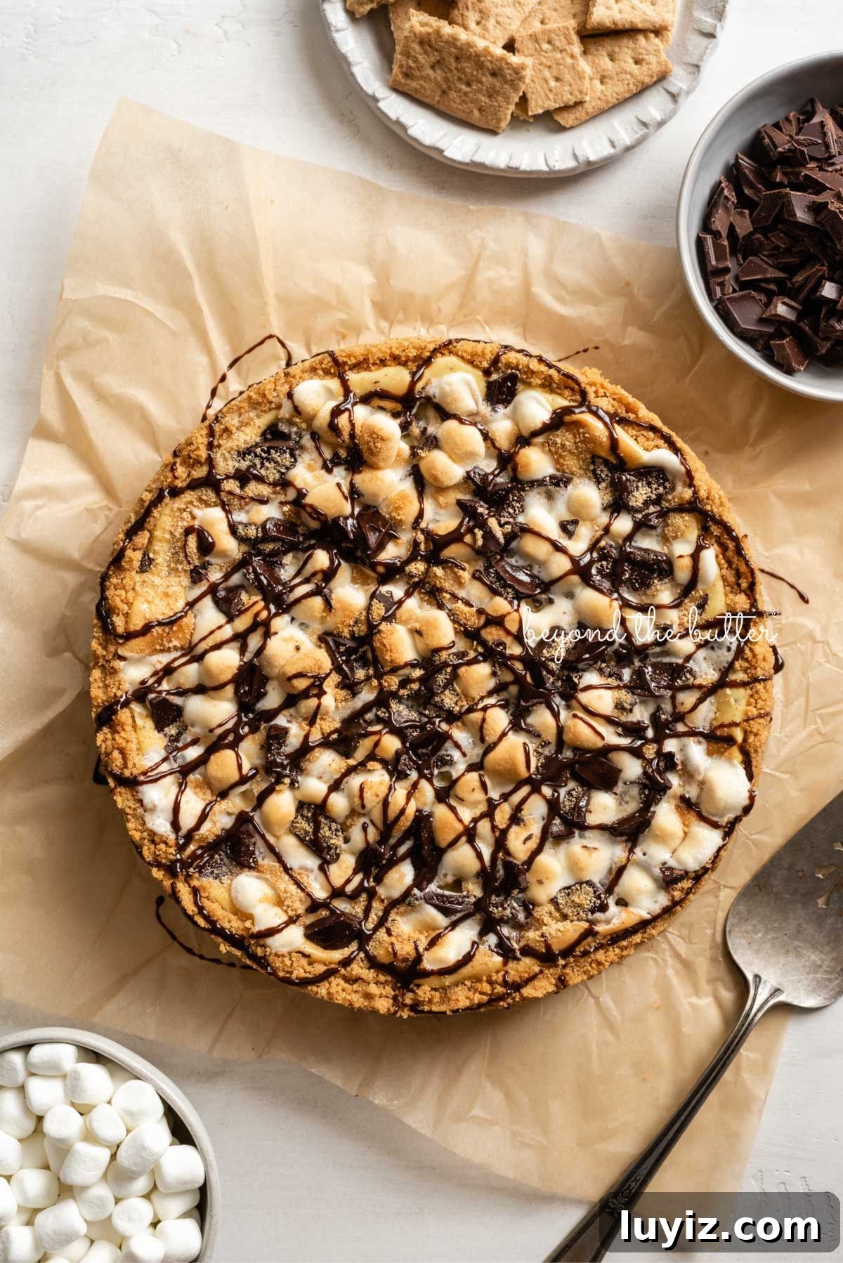 S’mores Cheesecake Recipe: Gooey Chocolate, Graham & Toasted Marshmallow 4 S'mores cheesecake topped with drizzled chocolate syrup placed on a sheet of brown parchment paper with a bowl of mini marshmallows, graham crackers, chocolate, and a cake server placed around it.