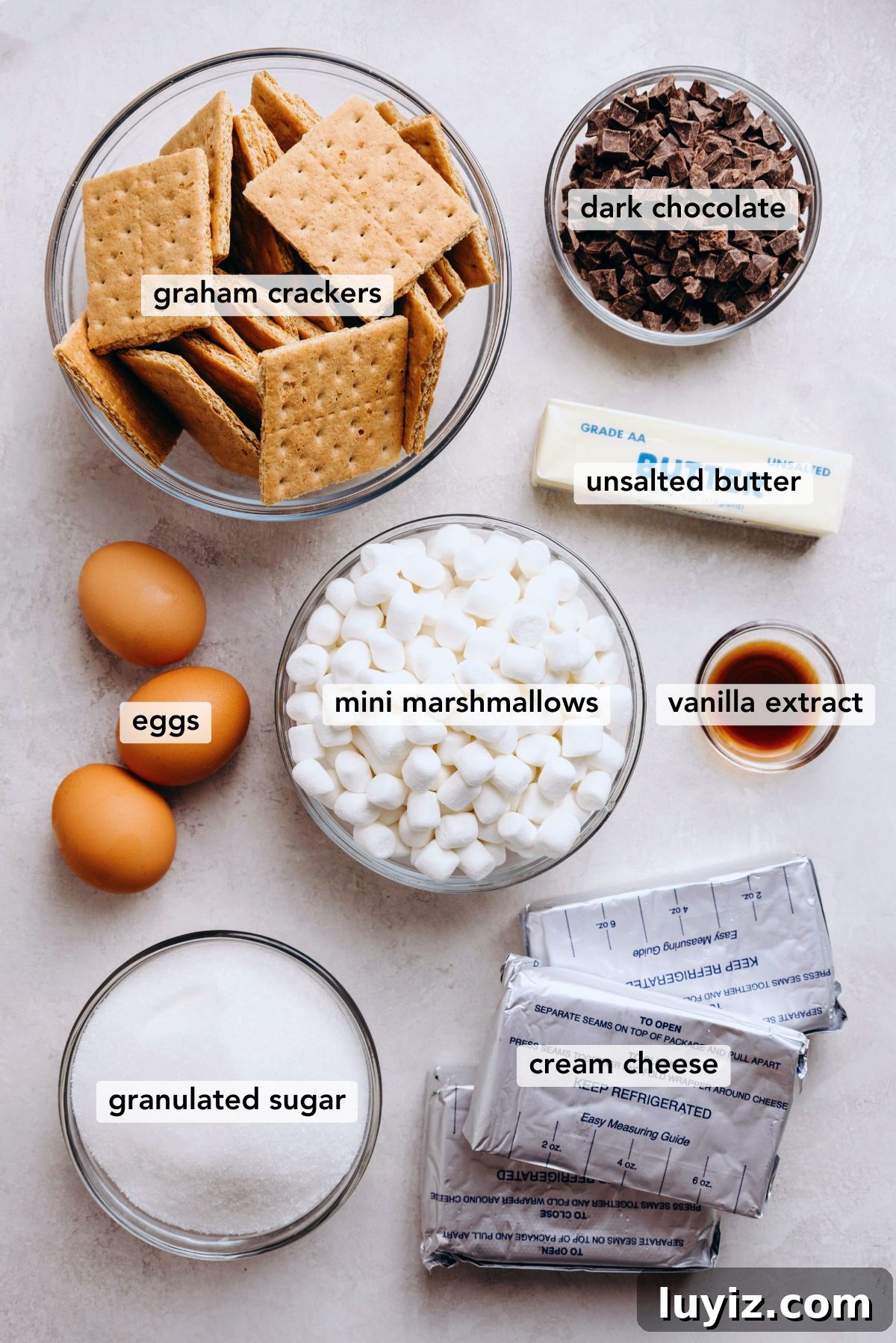 S’mores Cheesecake Recipe: Gooey Chocolate, Graham & Toasted Marshmallow 5 Ingredients in various size bowls labeled as graham crackers, dark chocolate, eggs, mini marshmallows, unsalted butter, vanilla extract, granulated sugar, and cream cheese on a white marbled background.