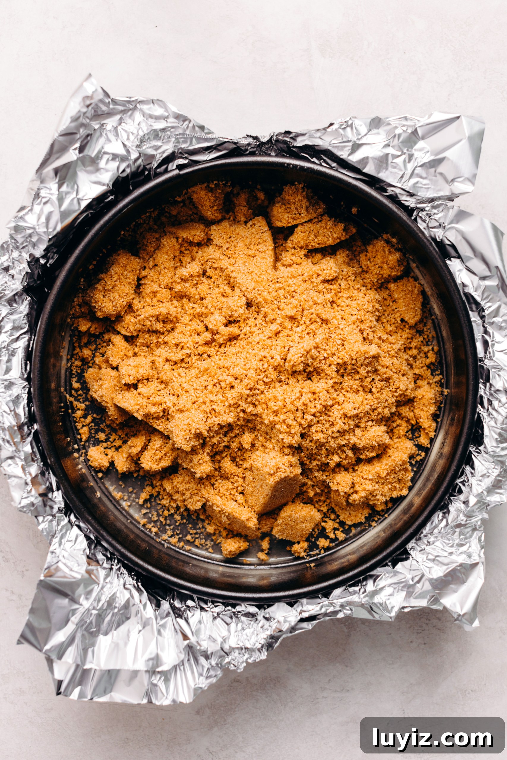 S’mores Cheesecake Recipe: Gooey Chocolate, Graham & Toasted Marshmallow 8 Graham cracker crust mixture placed on a aluminum foil wrapped springform pan on a textured white background.