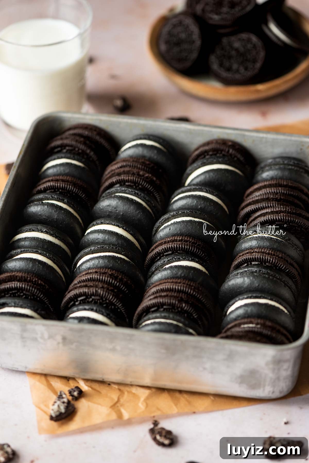 Oreo Cookie Macarons: Step-by-Step Recipe for Perfect Bites 3 Oreo macarons and oreo cookies in a vintage baking tin on parchment paper with a glass of milk and bowl of oreo cookies in the background.